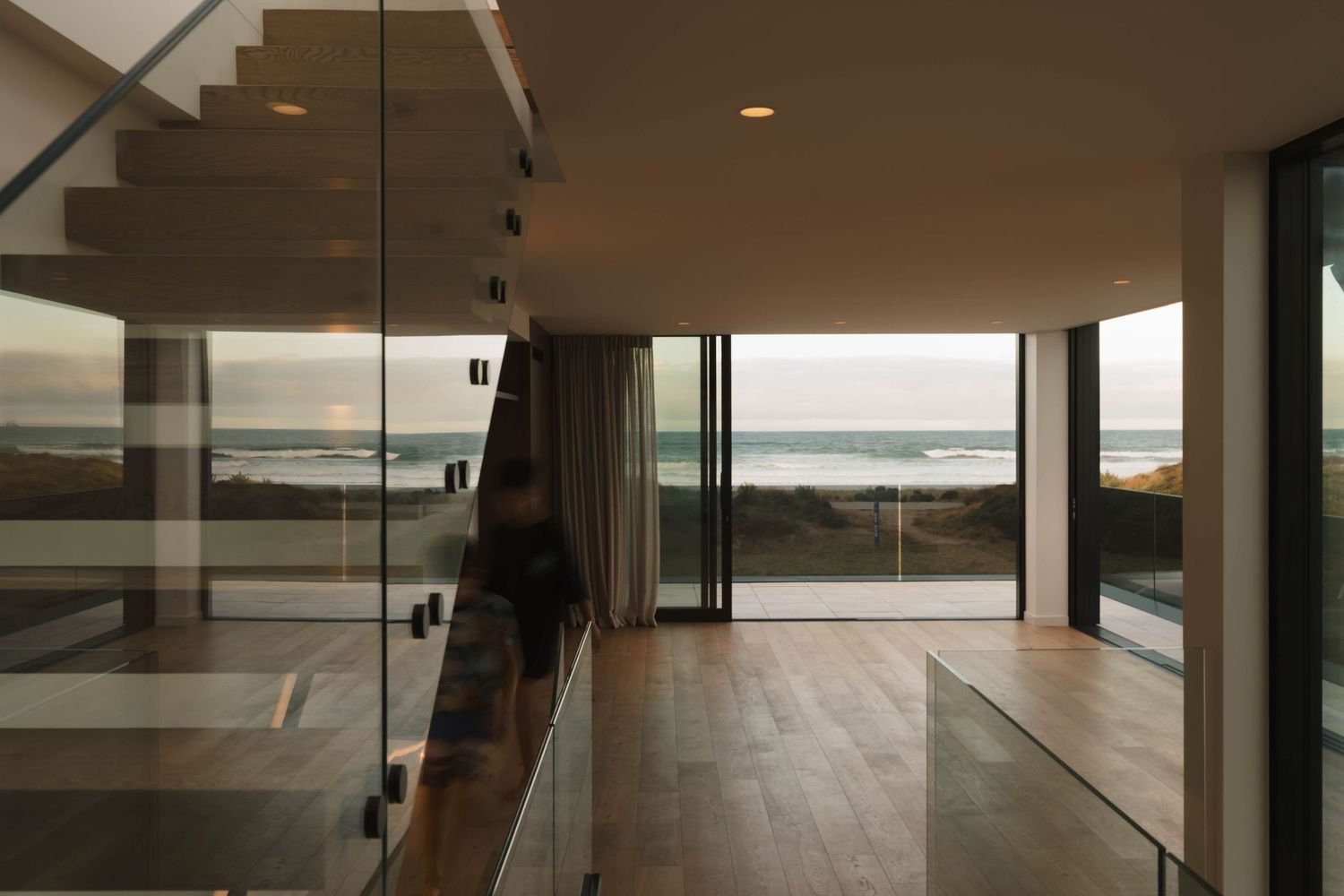 Contemporary coastal home interior with glass staircase and ocean view through floor-to-ceiling windows.