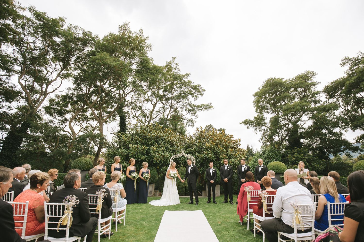 An outdoor garden wedding ceremony with guests seated on white chairs under tall trees during a late afternoon.