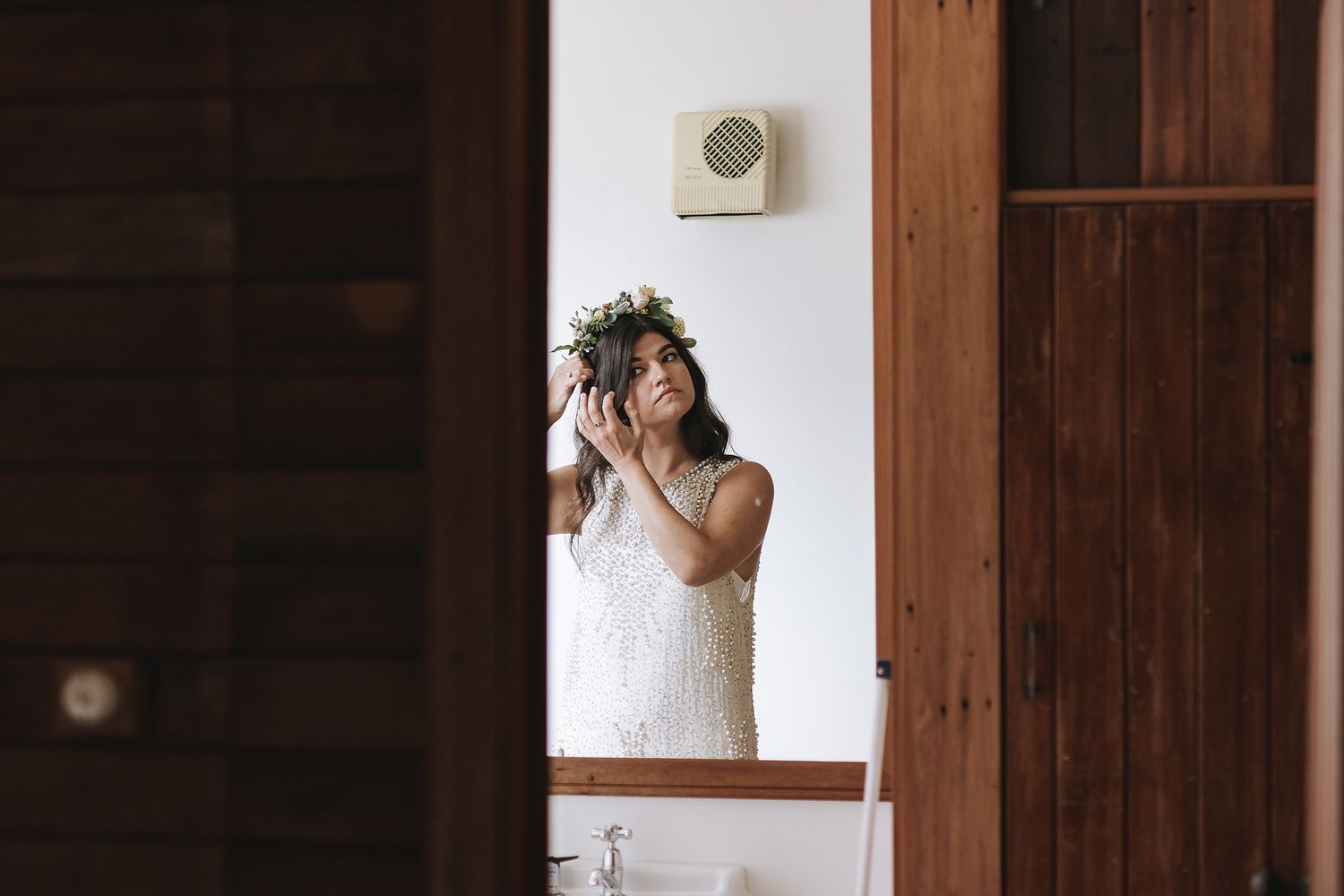 A figure wearing a white lace dress and floral crown stands in front of a mirror in a rustic wooden room.