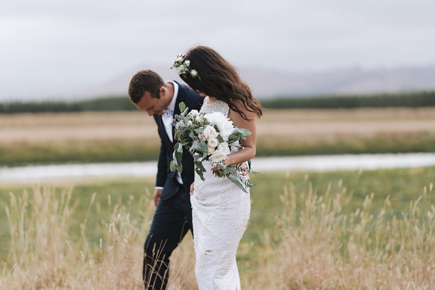 A couple in wedding attire embraces in a field of tall grass with autumn-colored foliage and mountains in background.