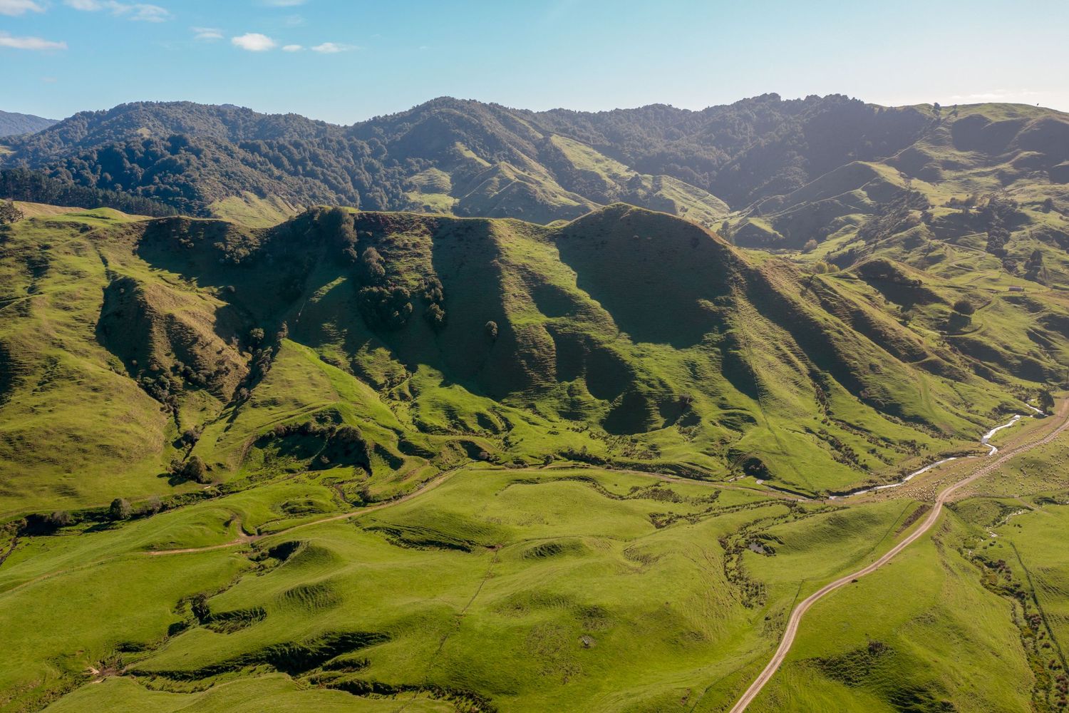 Winding road cutting through lush green mountain valleys with rolling hills and bright blue sky in the background.