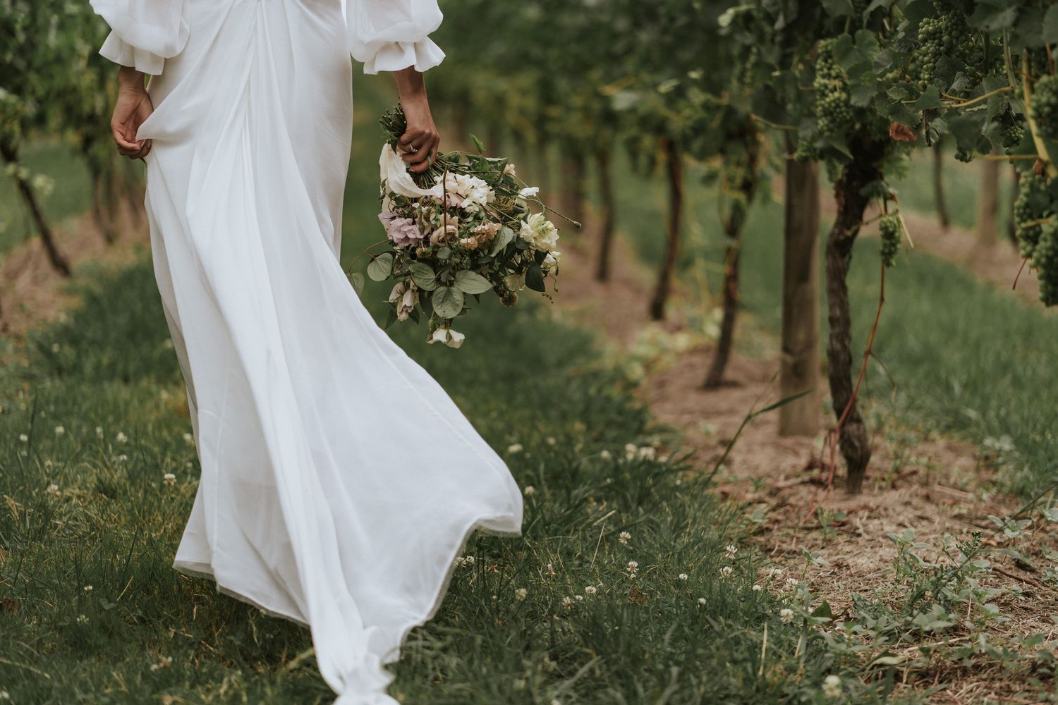 Flowing white wedding dress and bouquet photographed in a rustic vineyard setting during a summer wedding.