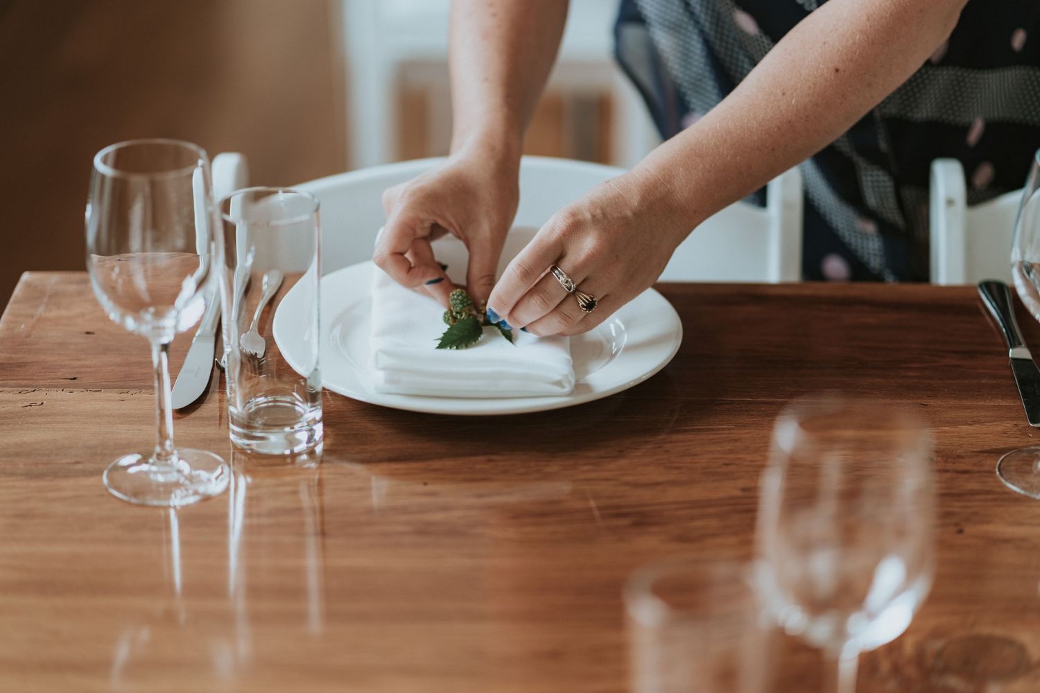 Person holding wine glasses at wooden dining table setting with plates and glassware arranged.