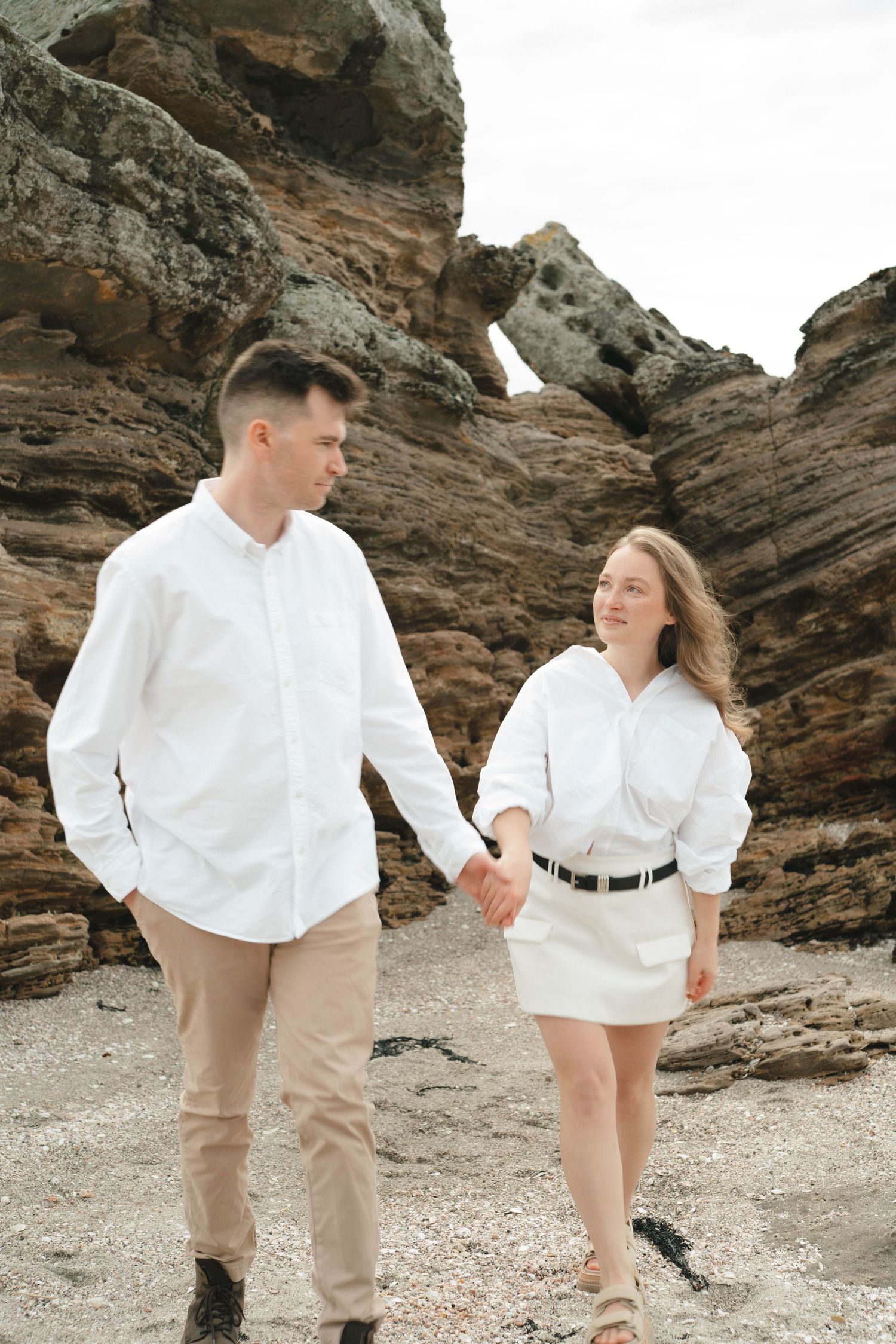 Playful couple in white clothing walking together among coastal rock formations.