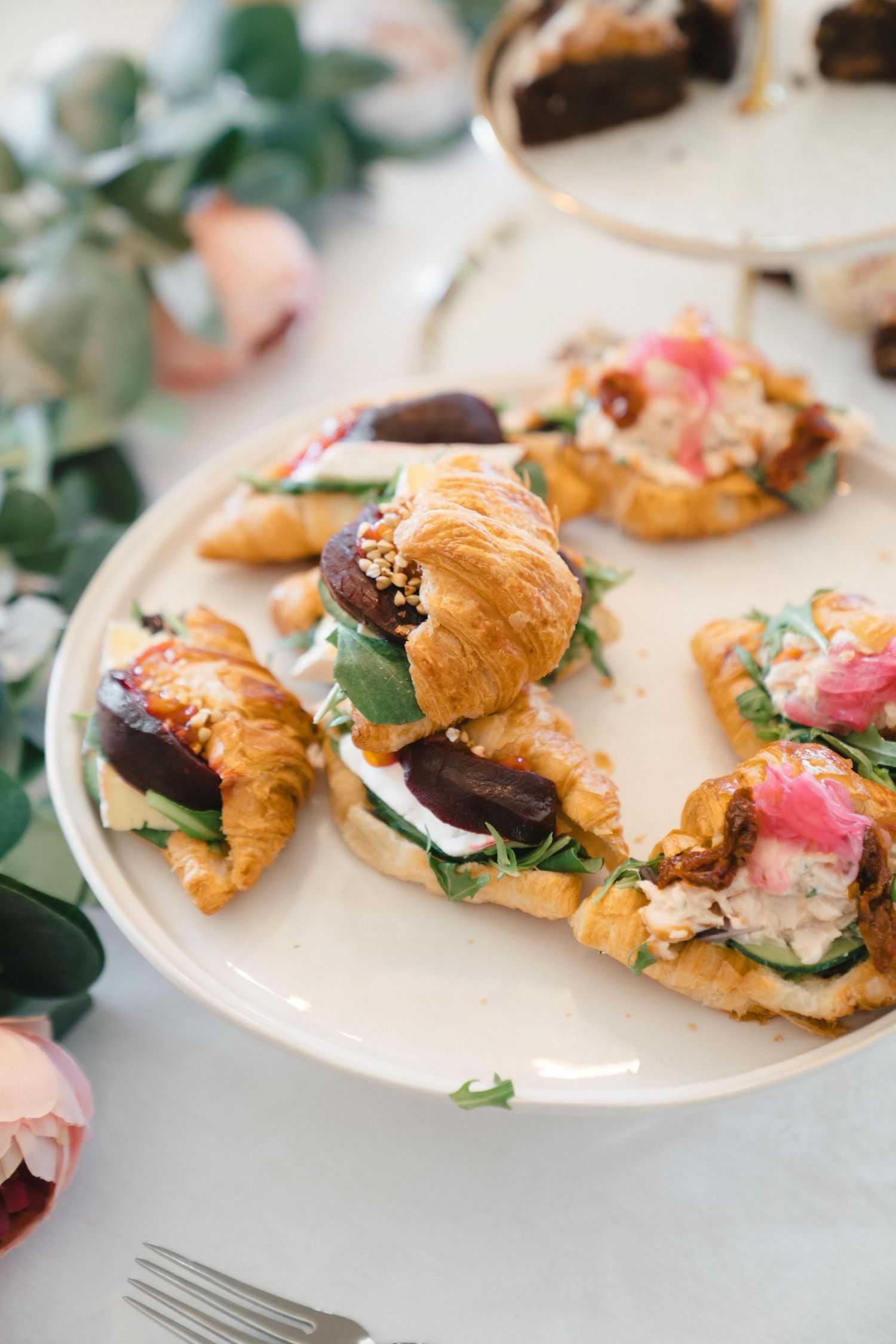 Elegant mini croissant sandwiches with colorful toppings served on a white plate with floral accents.