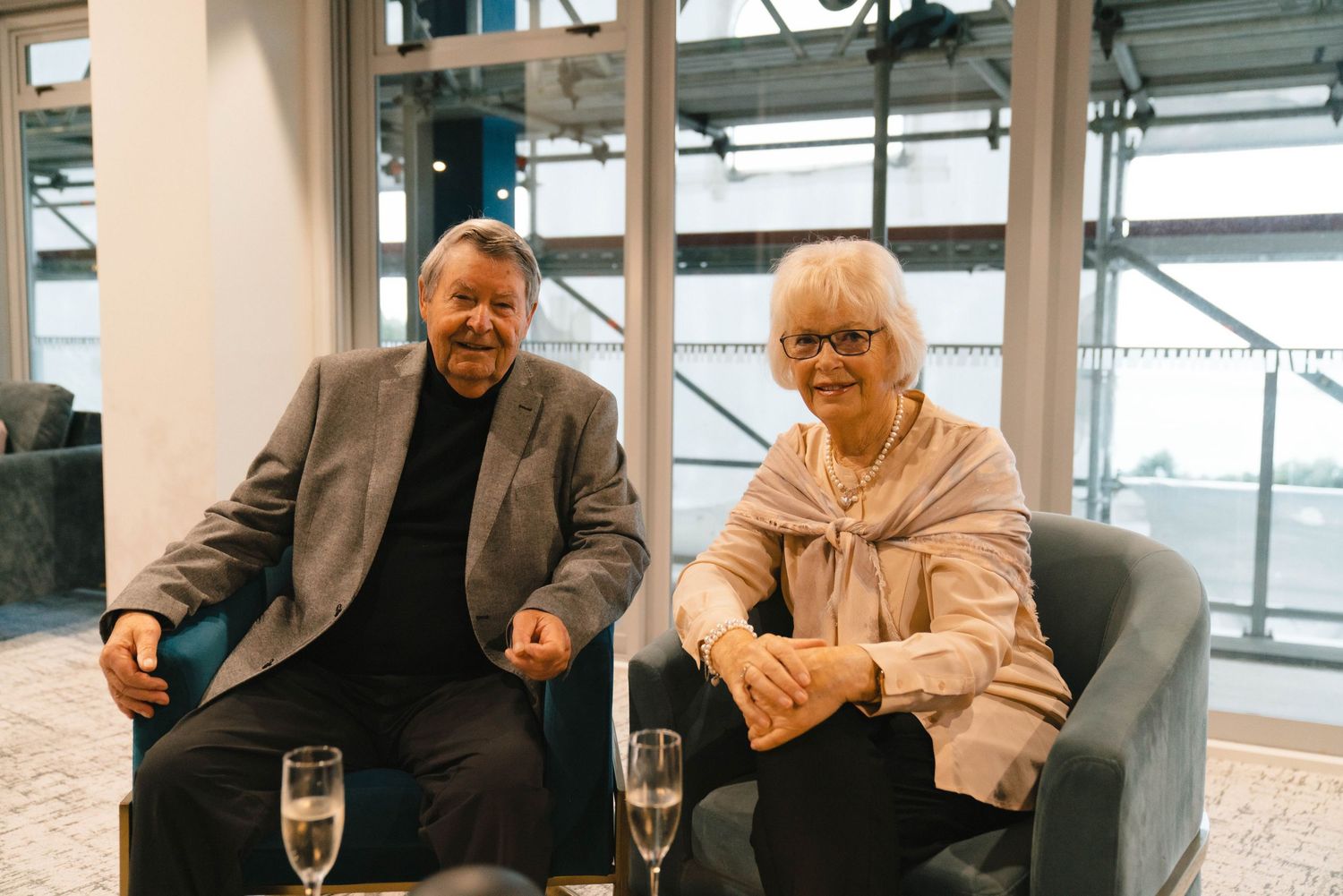 Two guests seated on gray chairs enjoying drinks at a social gathering.