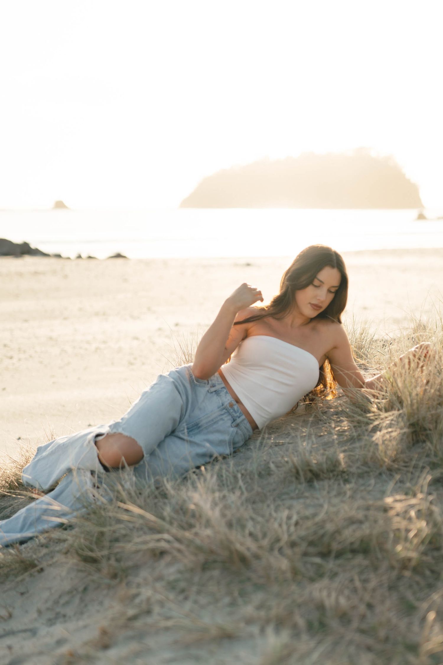 Woman in white top and denim reclines in coastal dune grass at sunset.
