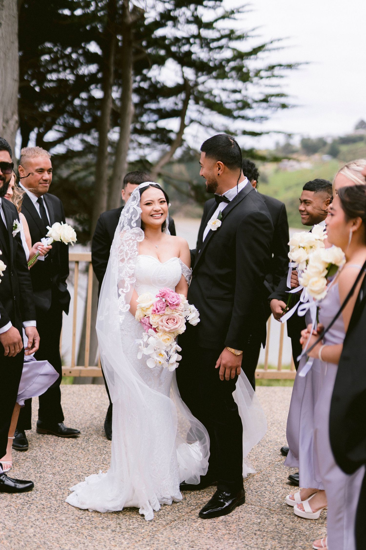 A wedding couple surrounded by their bridal party standing on a stone path with trees in the background.
