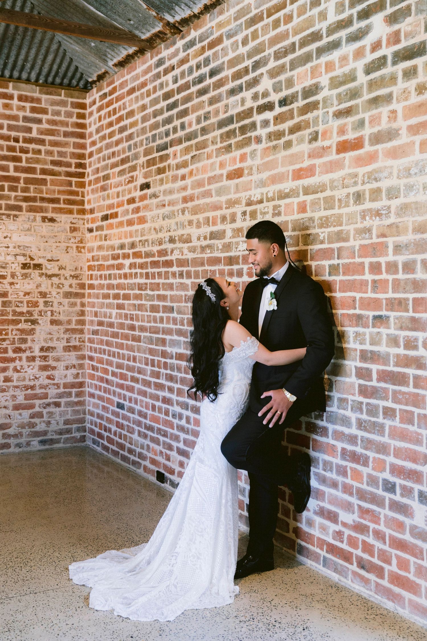 A newly married couple embraces while sitting on the floor against a rustic exposed brick wall.