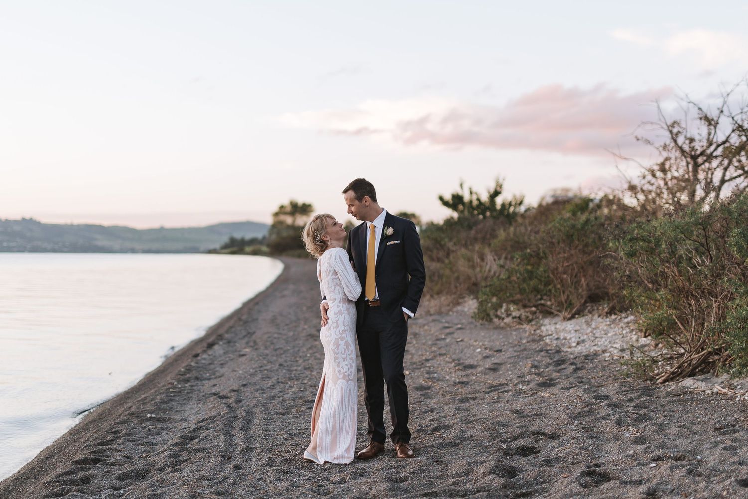 Newlywed couple embraces while walking along a scenic beach path at sunset with water and trees in background.
