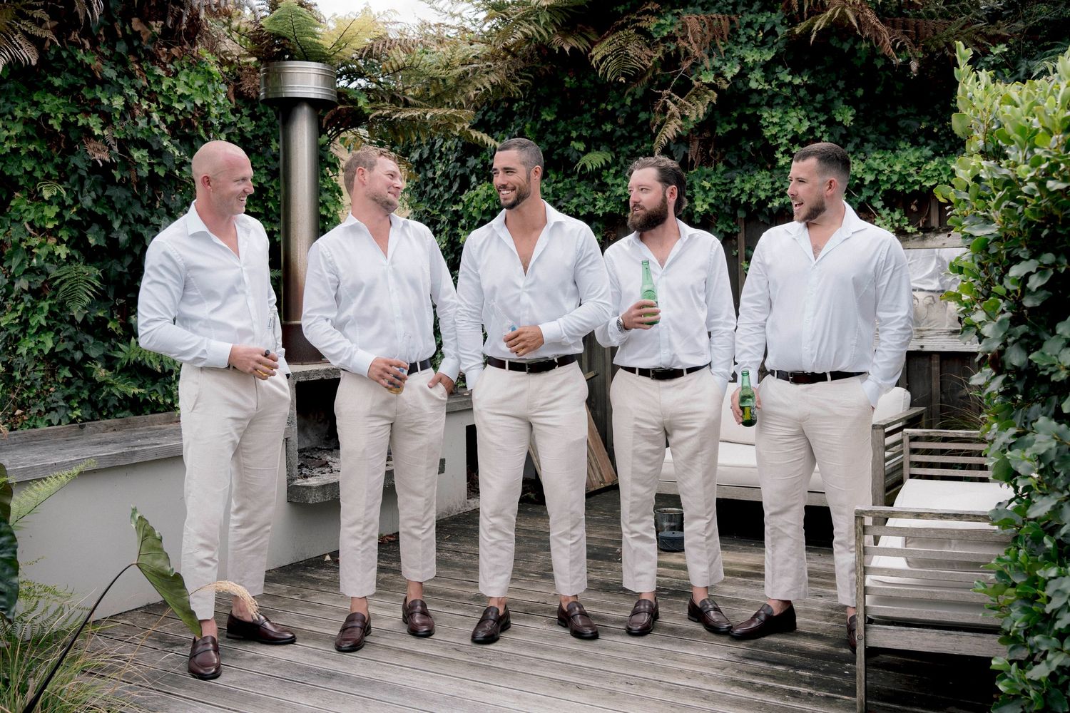 Five men in matching white casual attire and brown loafers stand together in a garden setting for a summer wedding photo.
