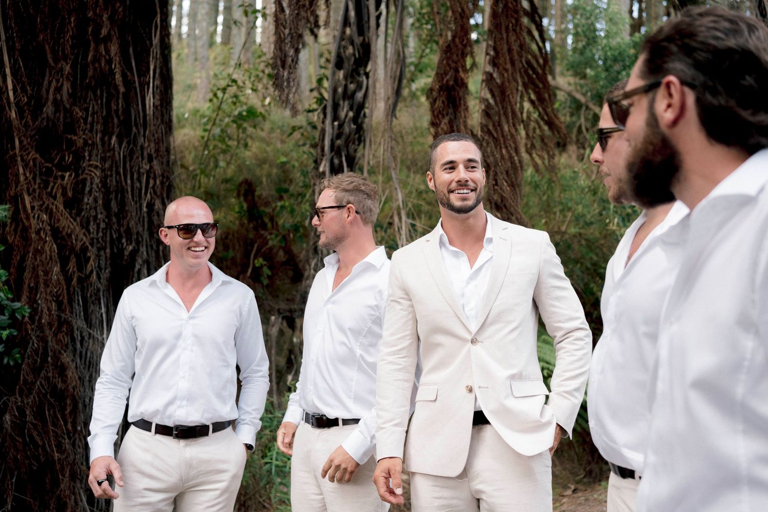 Group of people wearing white and cream colored formal attire gathered among tall redwood trees.