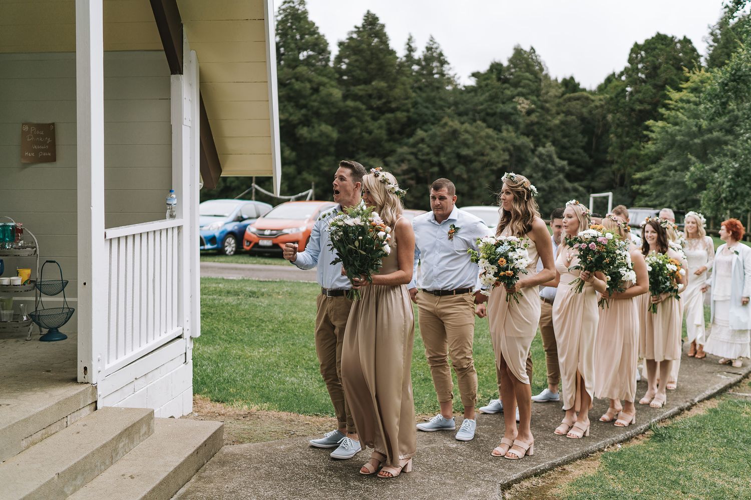 A wedding party in champagne-colored dresses arranges themselves in a line on a green lawn outside a white building.