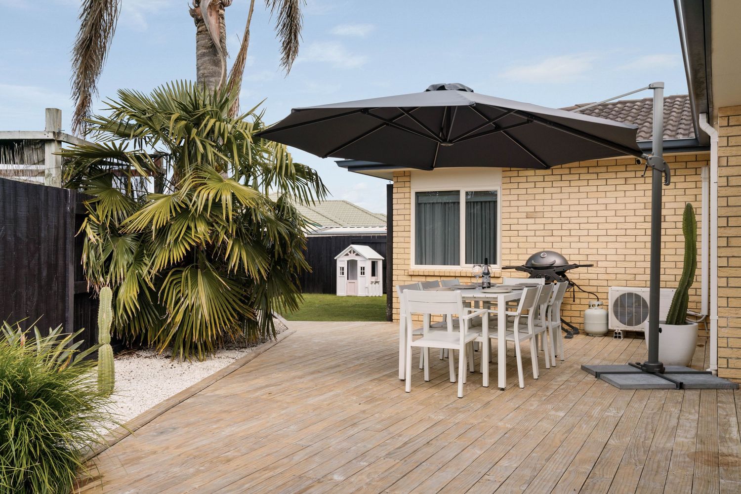 Outdoor patio with wooden deck, white dining set, black umbrella and tropical palm tree in backyard.