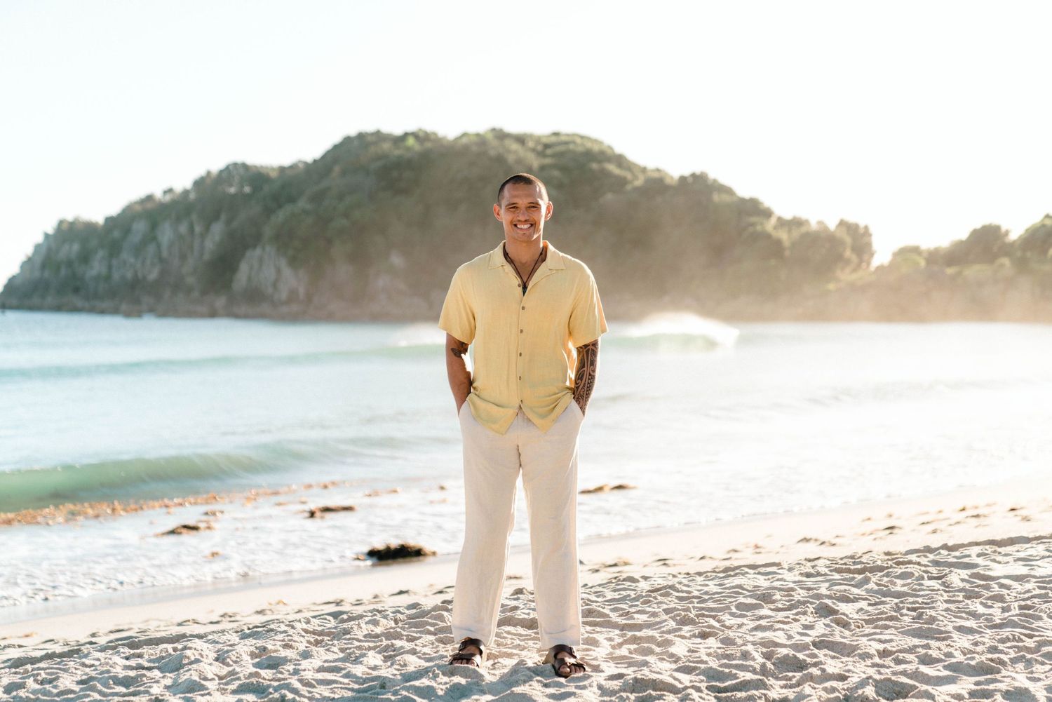 Person in casual yellow button-down and white pants poses on sandy beach at sunset with ocean view.