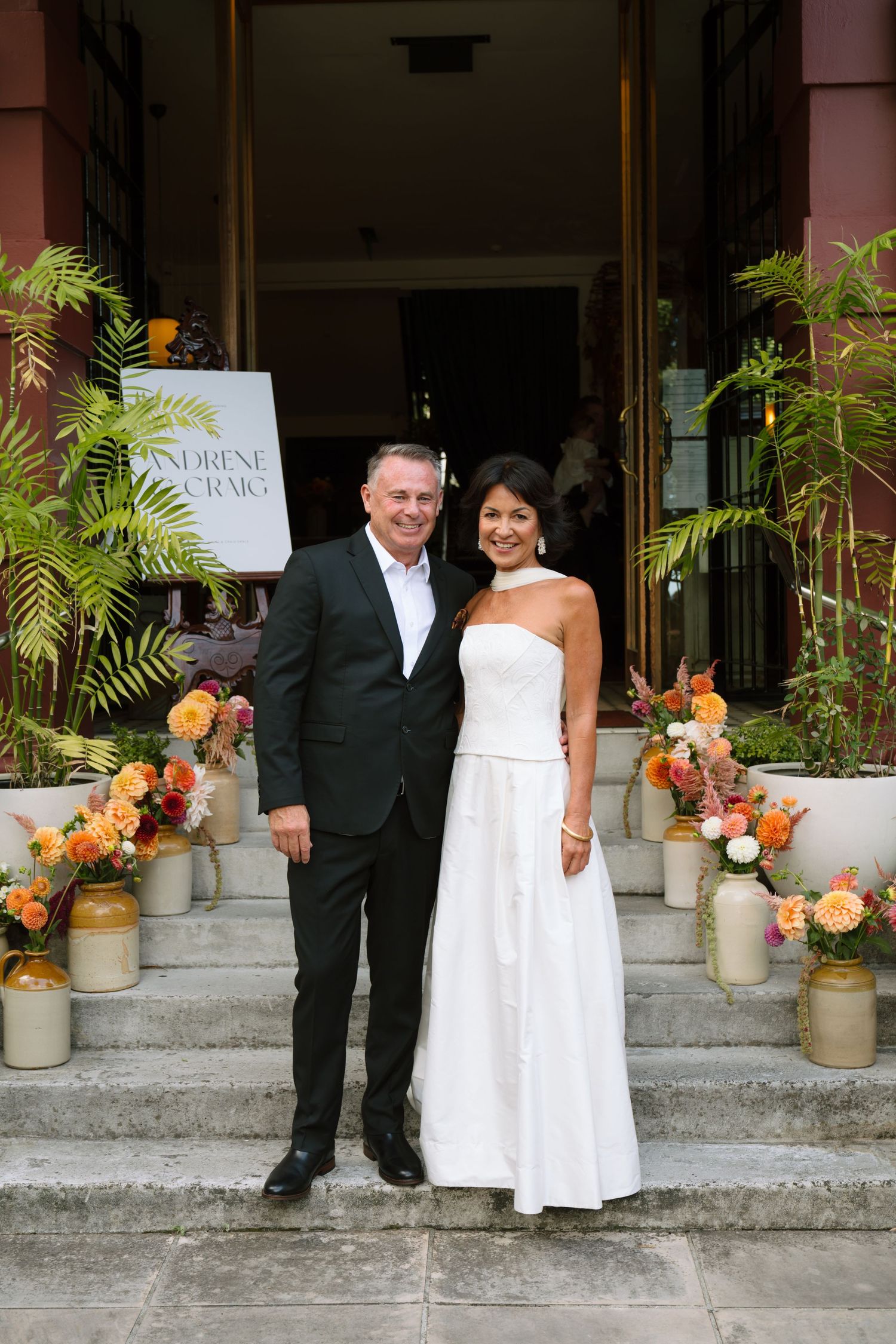 Couple in formal attire posing on steps adorned with orange floral arrangements in ceramic jugs outside a building. Bryony at Newfound.