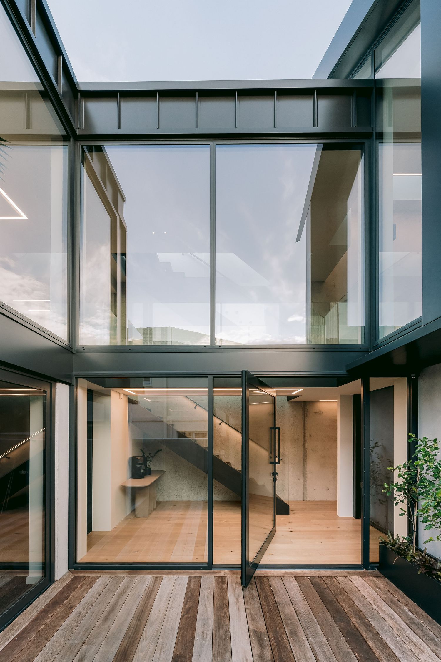 Modern glass and steel building entrance with wooden deck, pivot door, and interior staircase visible through floor-to-ceiling windows.