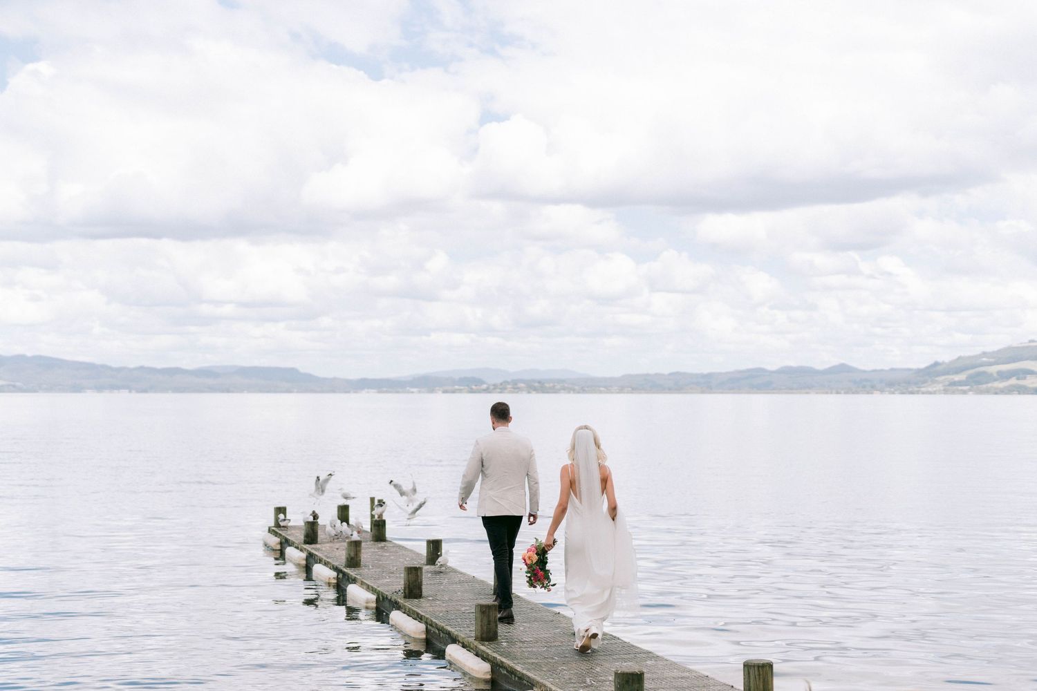 Couple walks hand in hand on wooden pier overlooking serene lake and mountain landscape.