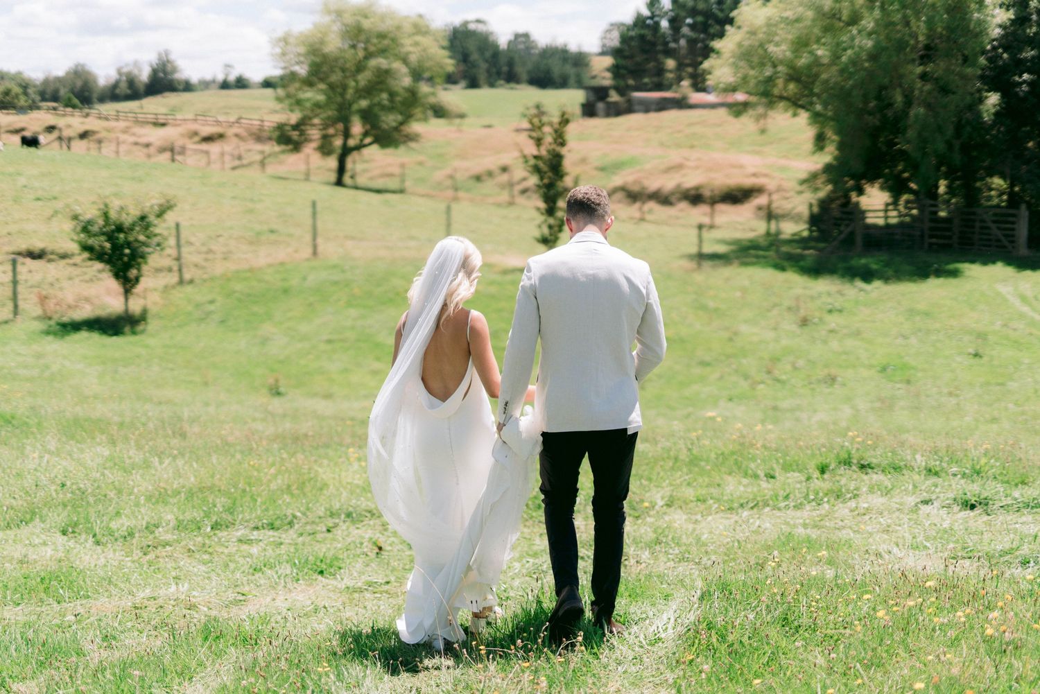 Couple holds hands while walking through a sunny green meadow on their wedding day.