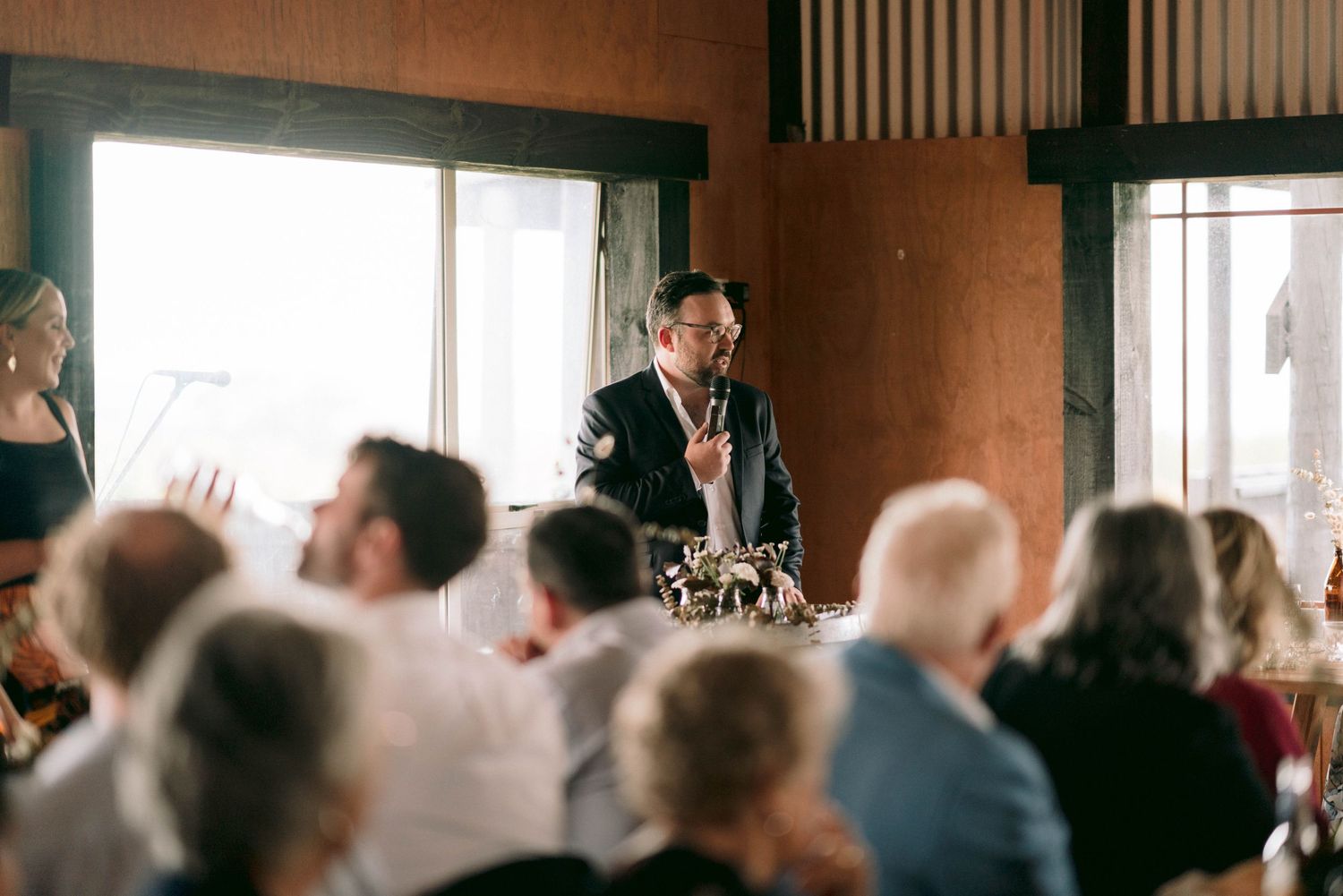 Speaker addresses seated audience in rustic indoor venue with natural lighting.