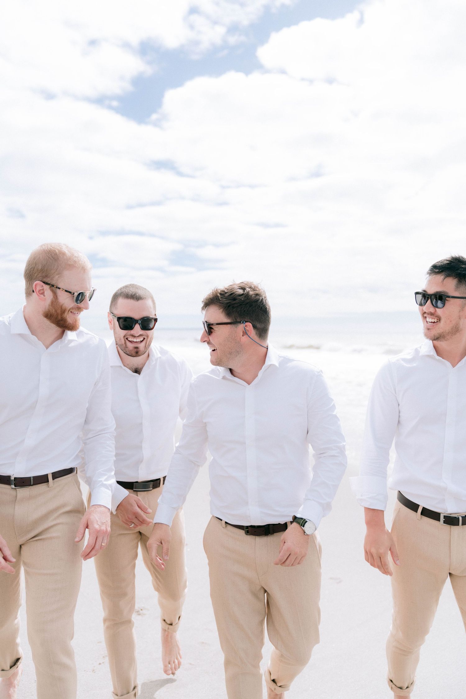 Groomsmen in matching white shirts and khaki pants walking along a beach on a sunny day.