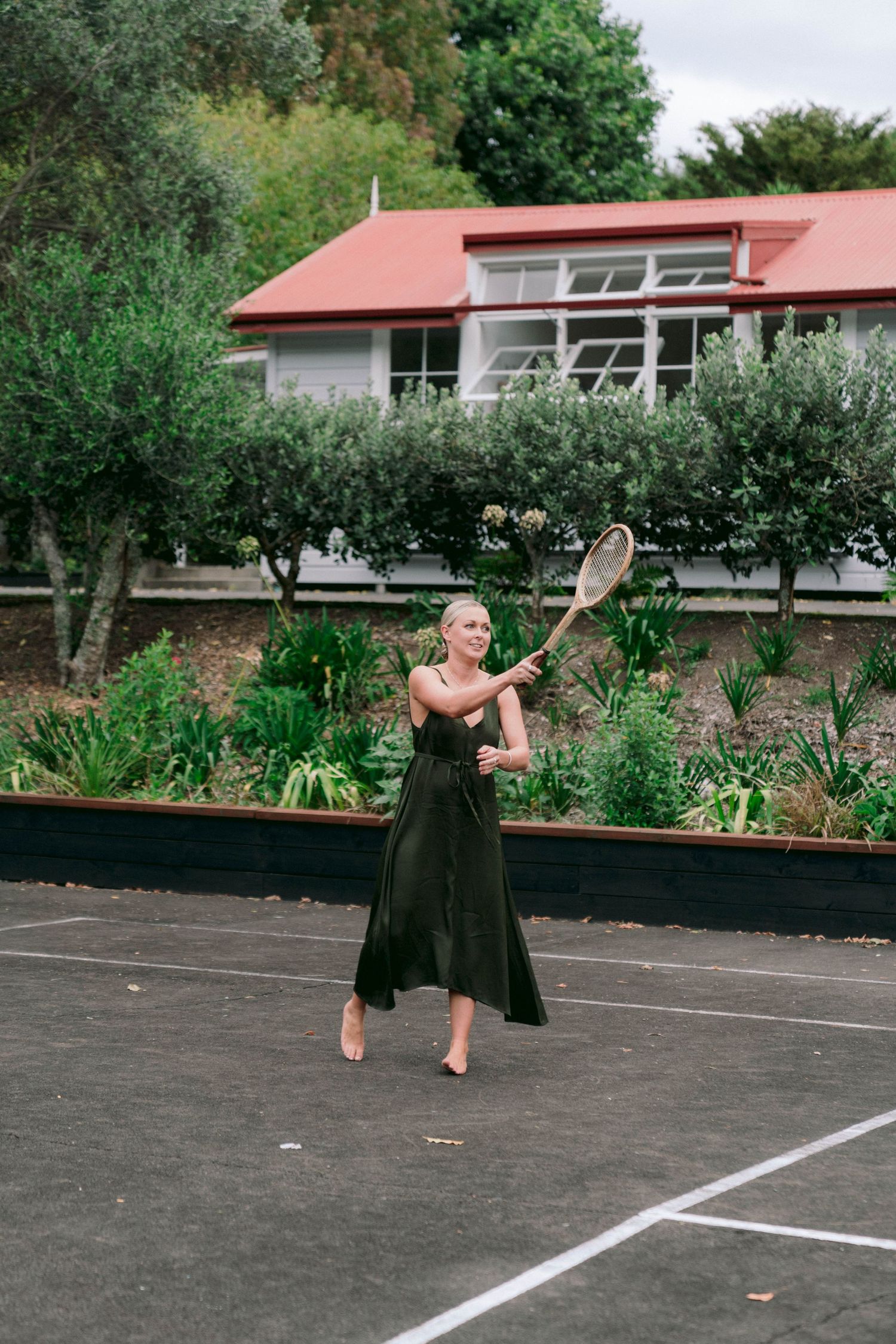 A person in a dark dress plays tennis on a court with a house and garden in the background.