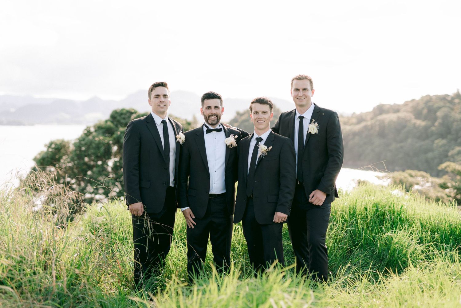 Groomsmen in black tuxedos standing together on scenic hillside with coastal views.