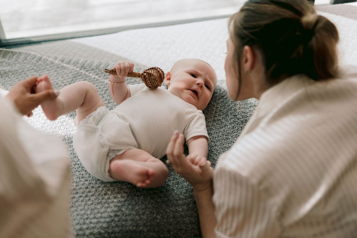 A person holds and plays with a newborn baby while resting together on a cozy gray blanket.