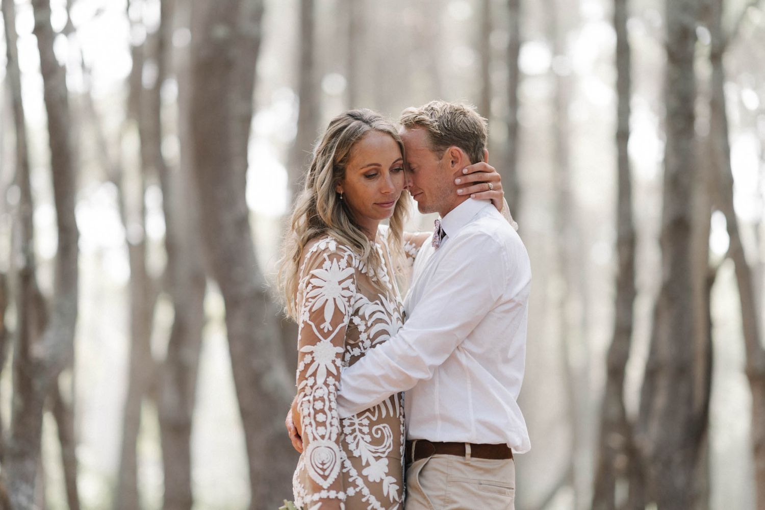 Romantic couple embraces in dreamy forest setting wearing white lace dress and casual button down shirt.