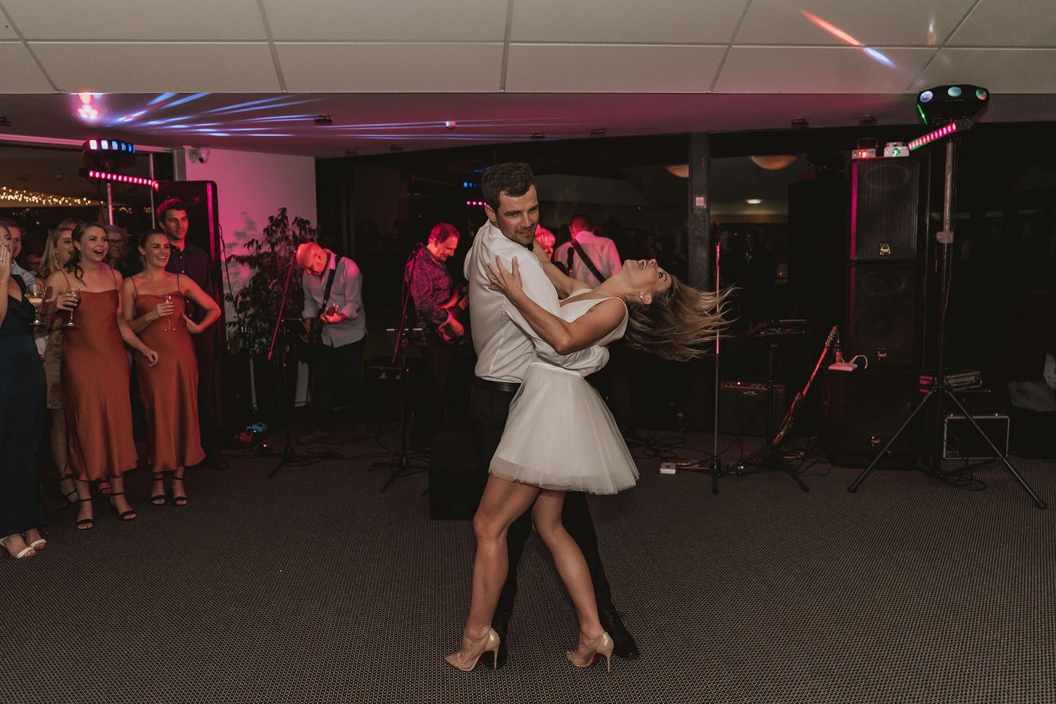 Dancing couples move across a dimly lit ballroom floor illuminated by pink and purple lighting during a social event.