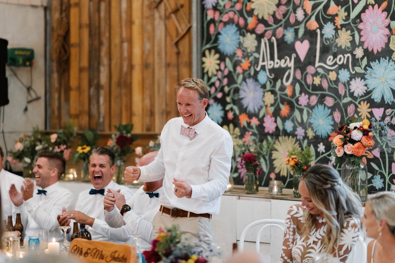 Reception guests in white attire enjoy dinner conversation at rustic barn wedding with colorful floral wall backdrop.