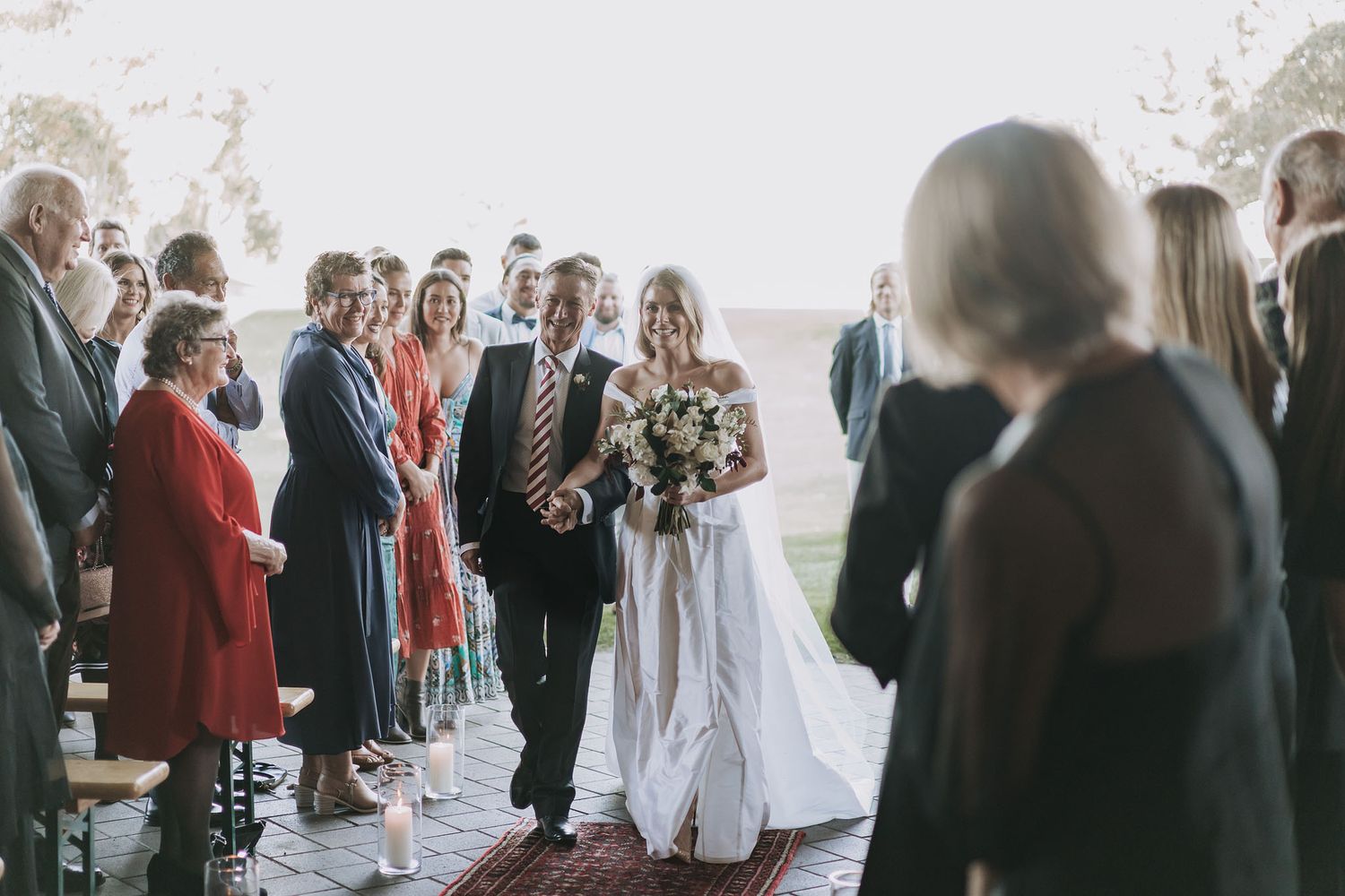 Wedding guests gather along elegant stone archways as the bride enters with her bouquet during an outdoor ceremony.