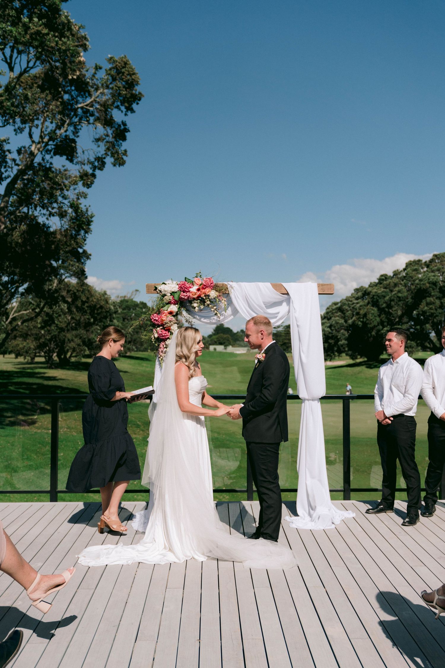 Outdoor wedding ceremony on wooden deck with white fabric arch and floral decorations.