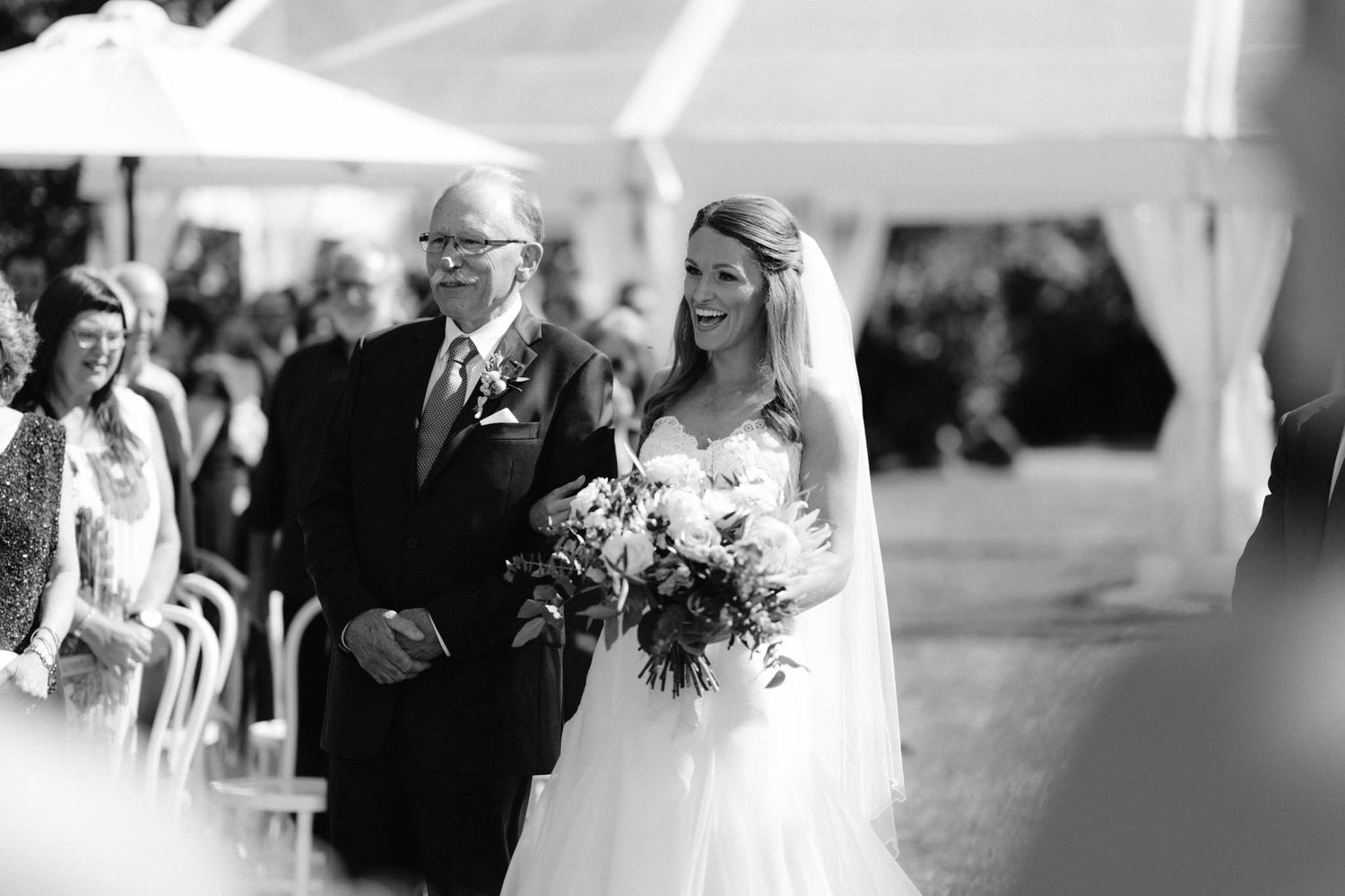 Black and white photo of a wedding ceremony processional with a bride walking down the aisle holding a bouquet.