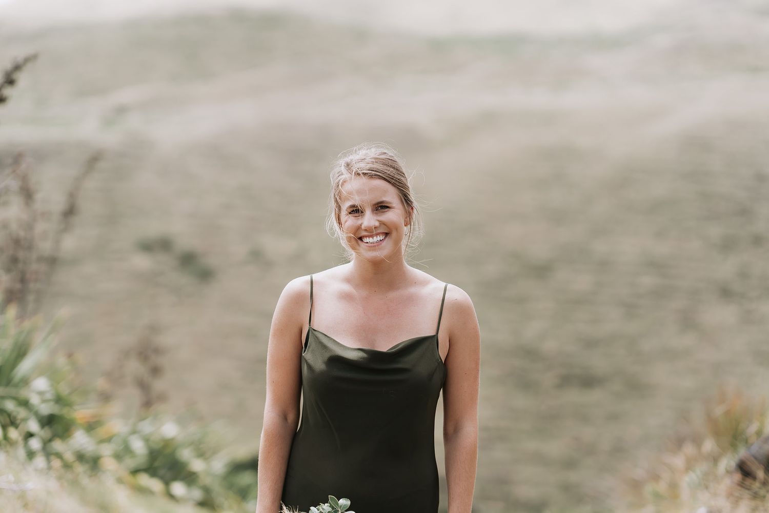 Person in a black sleeveless dress stands outdoors against a mountainous landscape with green hills.