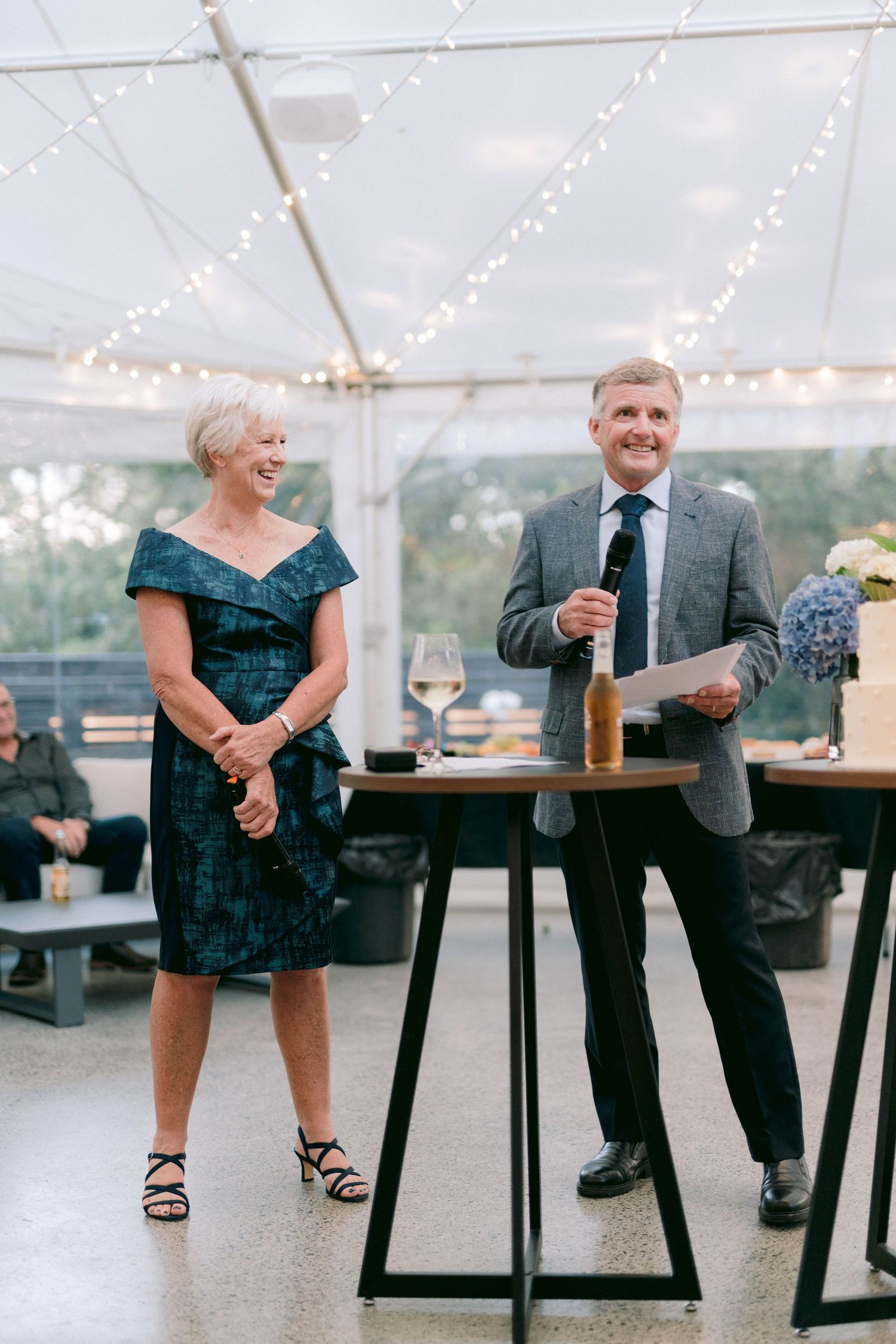 Two people stand at tall cocktail tables under twinkling string lights during an elegant event in a tent reception.