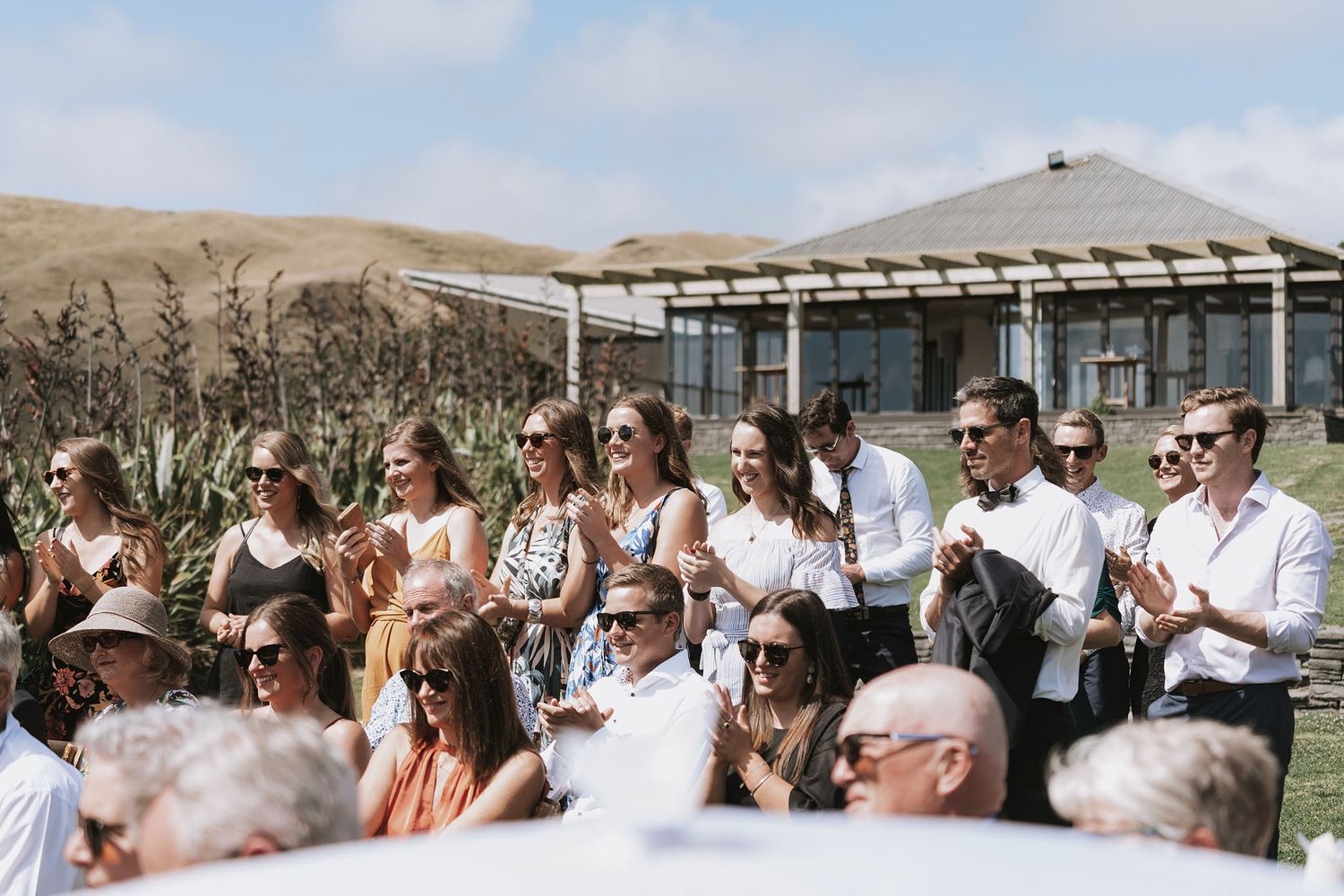 A crowd of spectators watches from a viewing area with a pergola structure at an outdoor event on rolling hills.