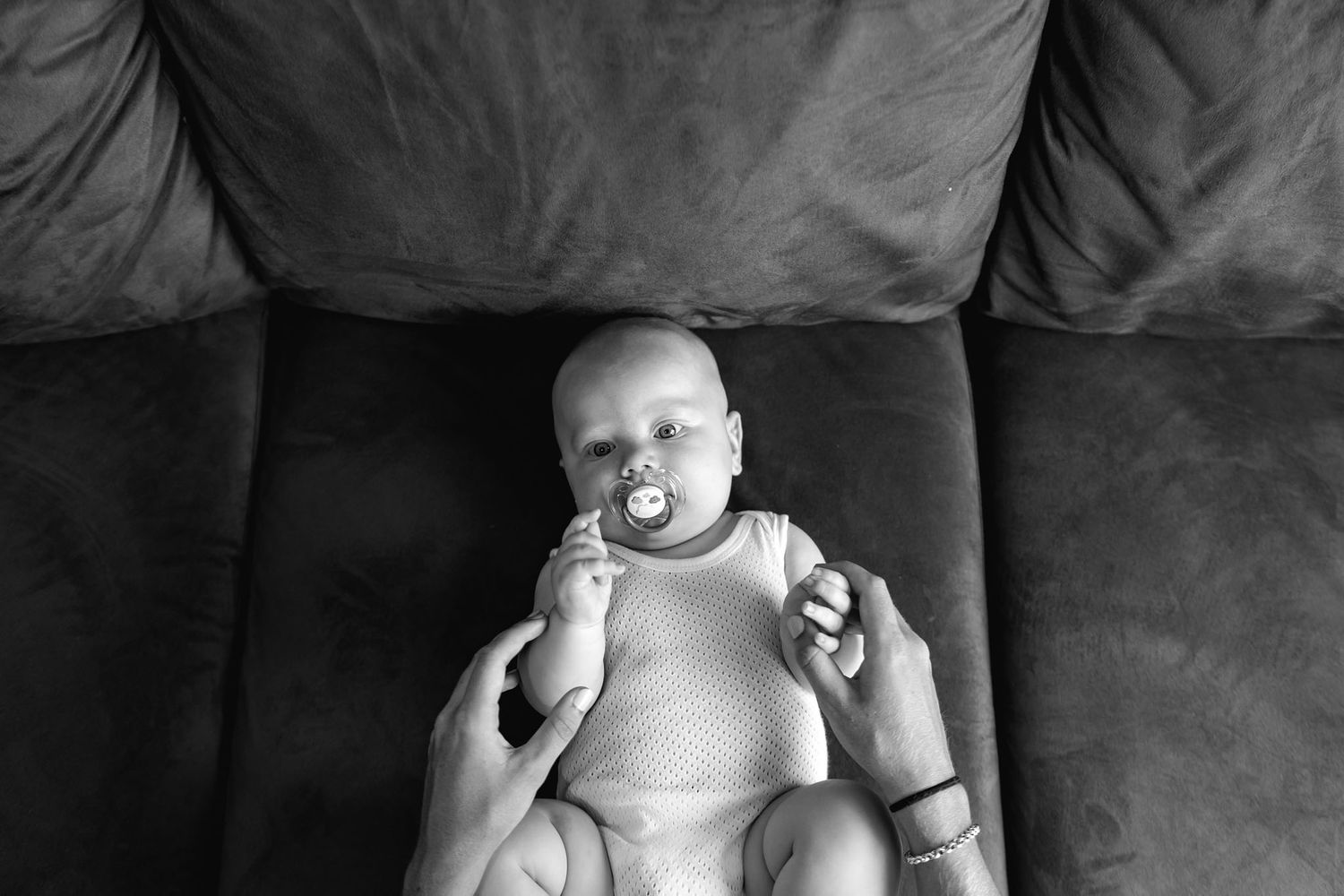 Black and white image of a baby in a diaper resting on a dark couch with a pacifier.