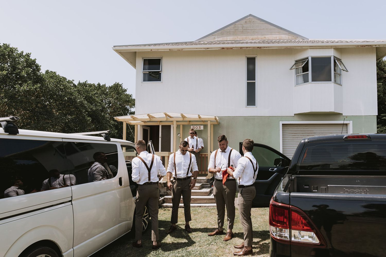 Groomsmen in suspenders and dress shirts gather between parked vehicles outside a white two-story house.