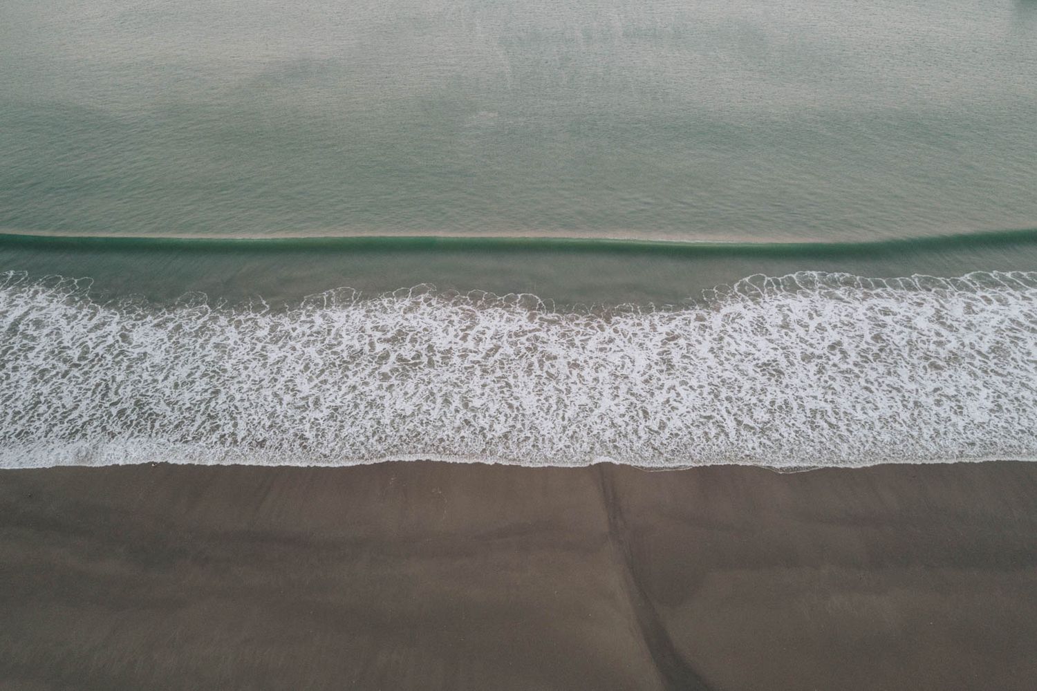 Aerial view showing a series of ocean waves rolling toward the beach with white foam breaking against the shore.