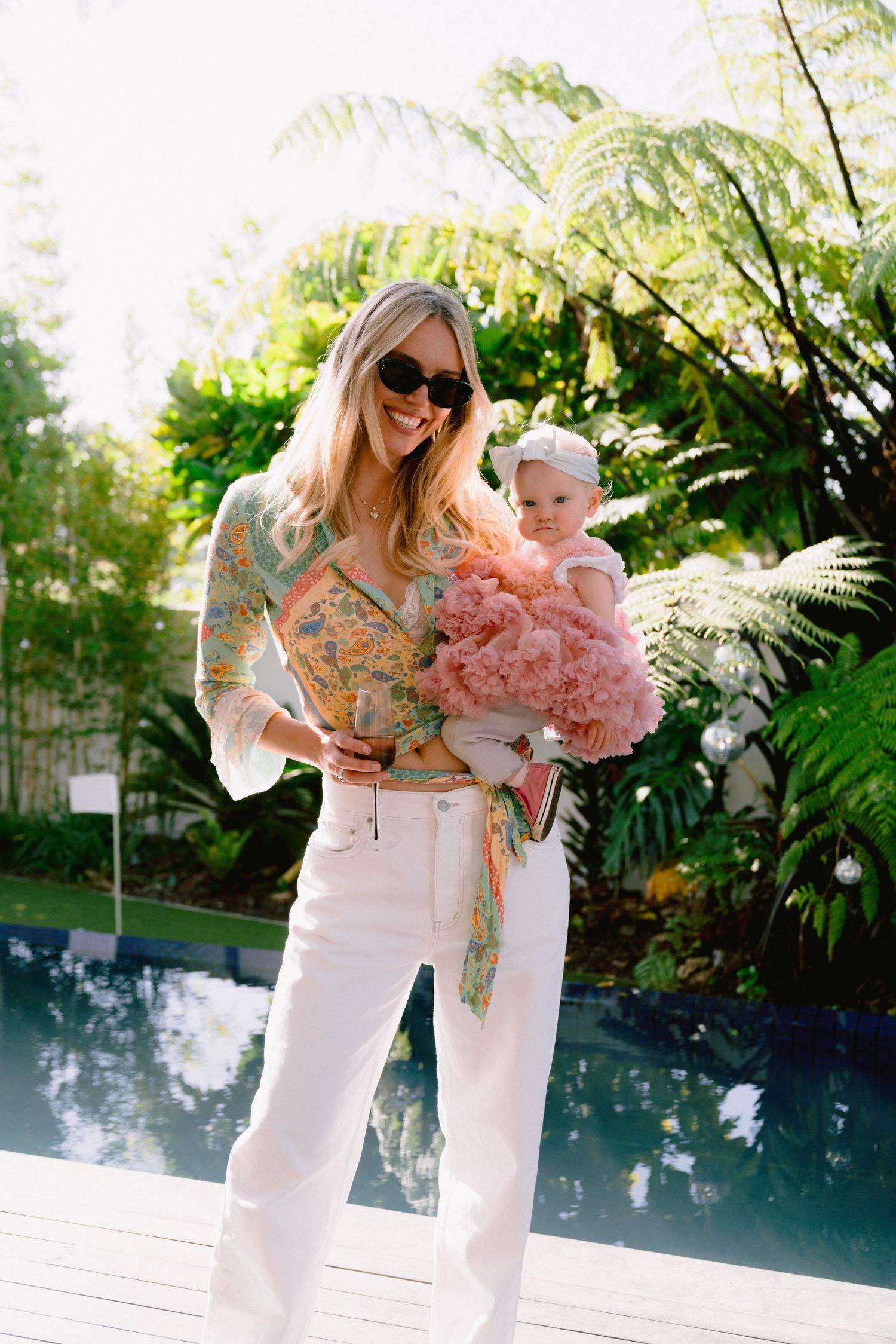 Woman in white dress holding a baby and pink flowers by a pool with tropical landscaping.
