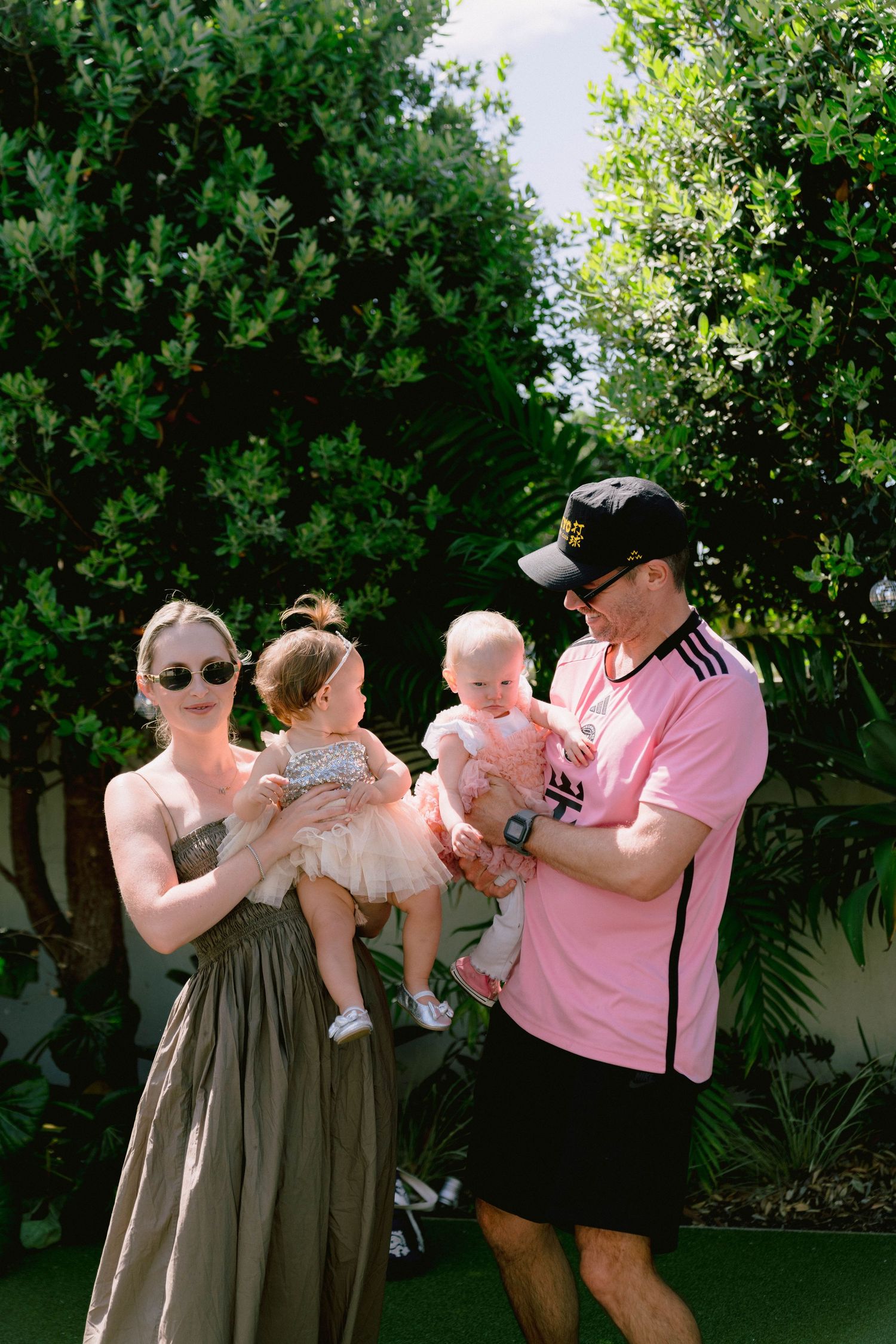 A family plays with their baby outdoors in a garden setting with lush green foliage.