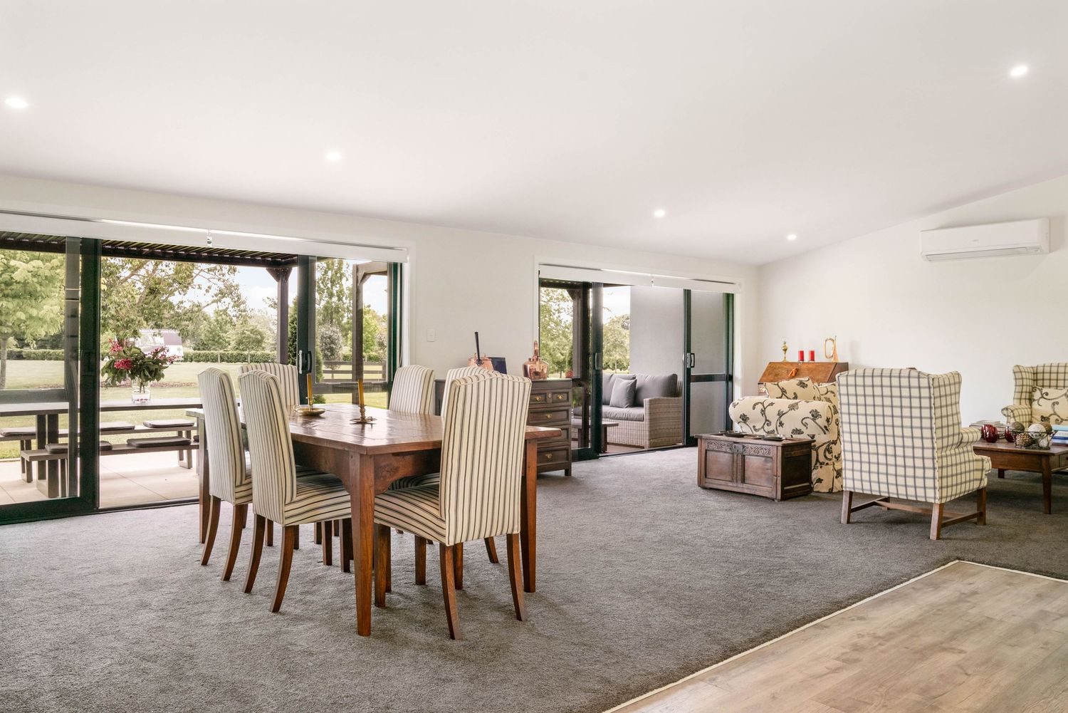 Modern open-plan living room with dining area features grey carpet, wooden furniture and large windows overlooking greenery.