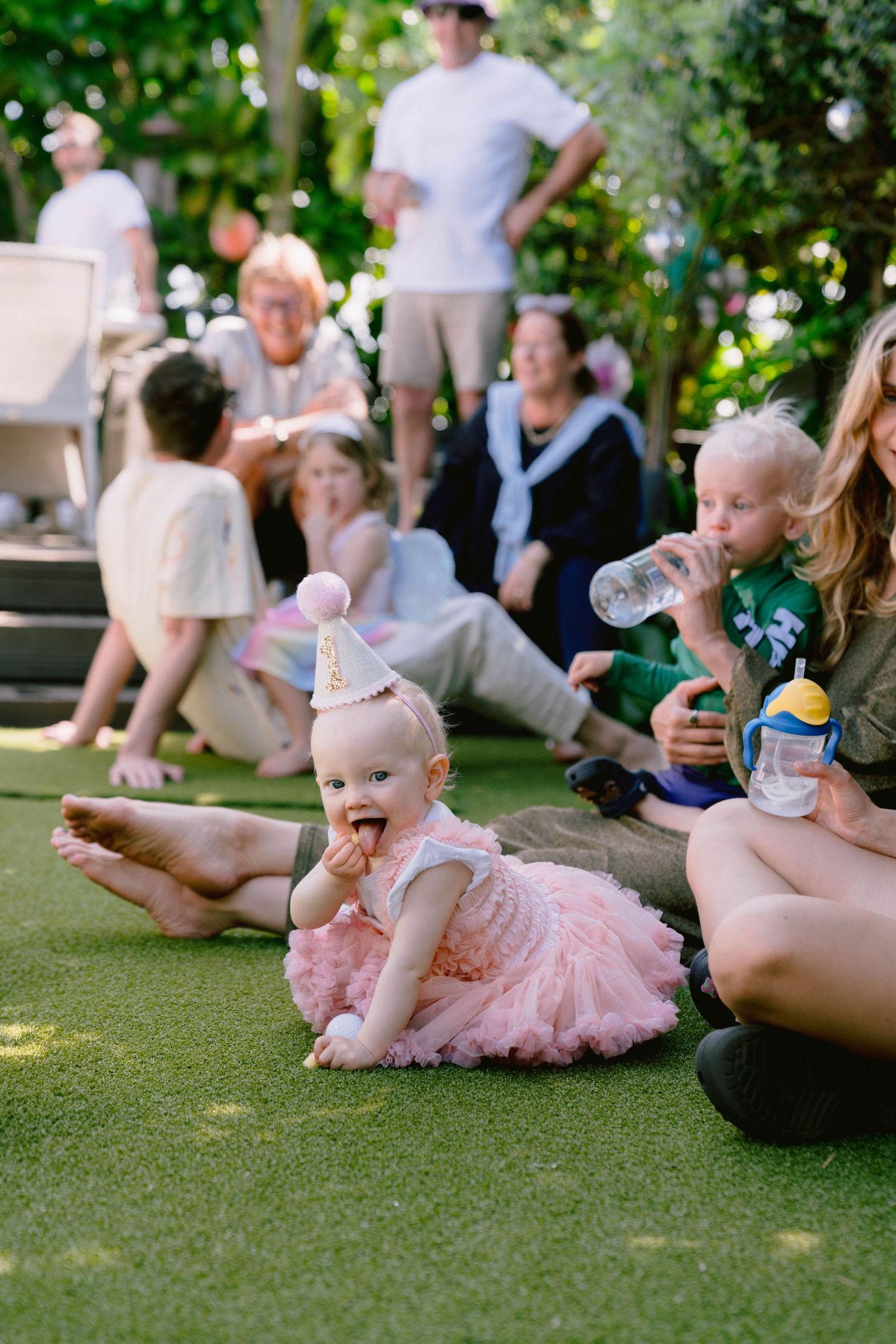 A baby in a pink tutu dress sits on grass during an outdoor gathering with other people in the background.