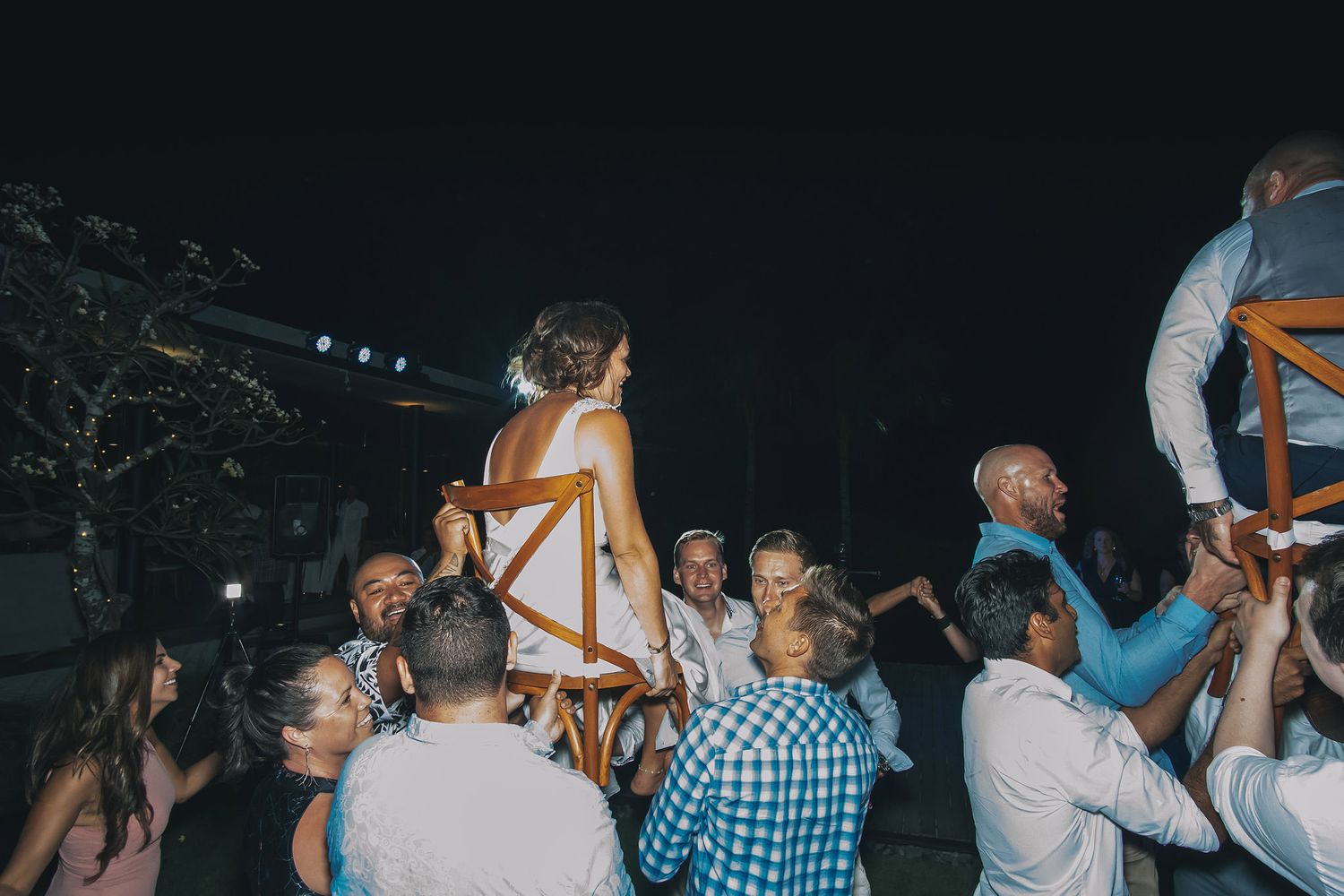 Nighttime wedding celebration with guests lifting people up on chairs during traditional reception dance.