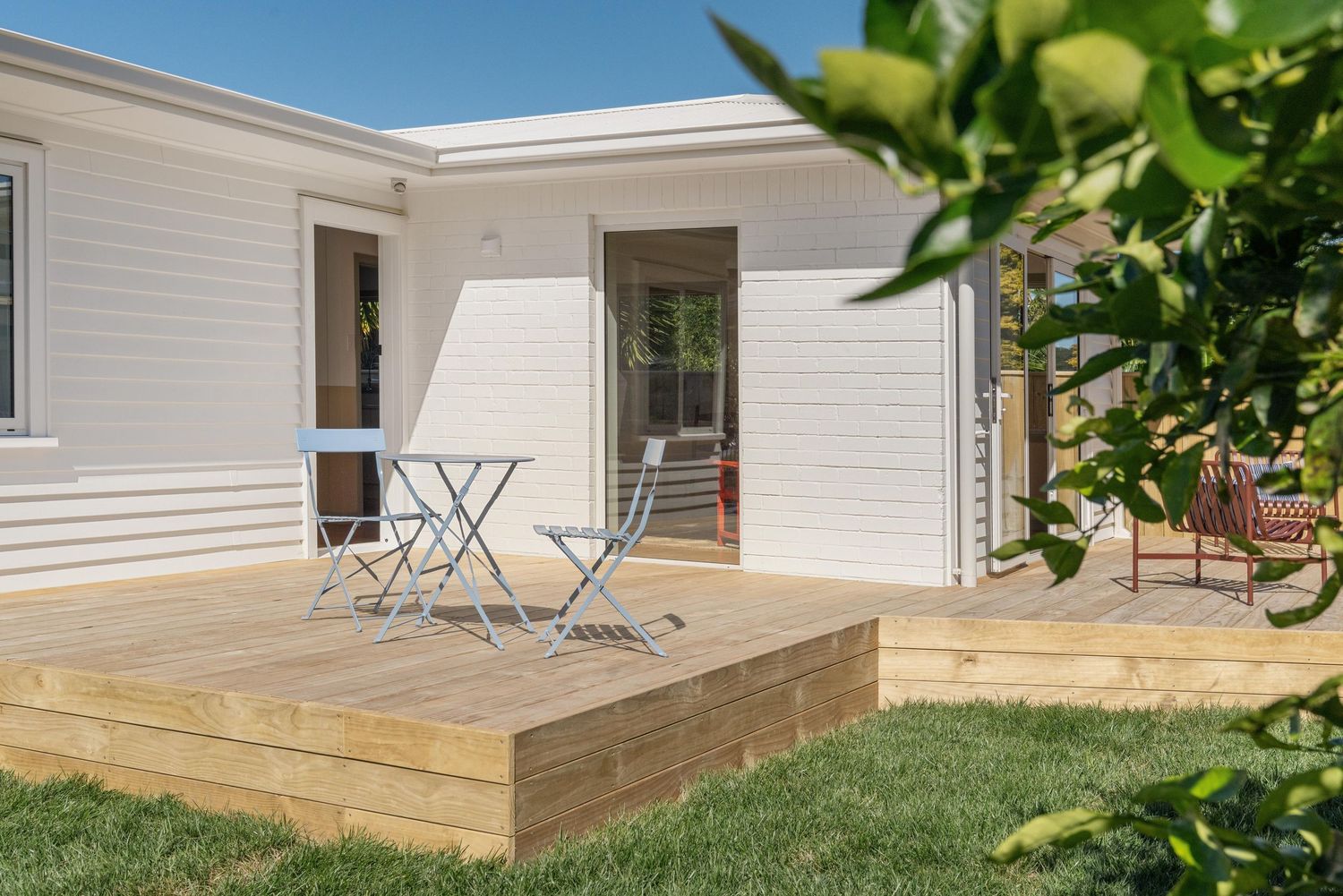 Modern white house exterior with wooden deck patio featuring folding chairs and green plants in foreground.