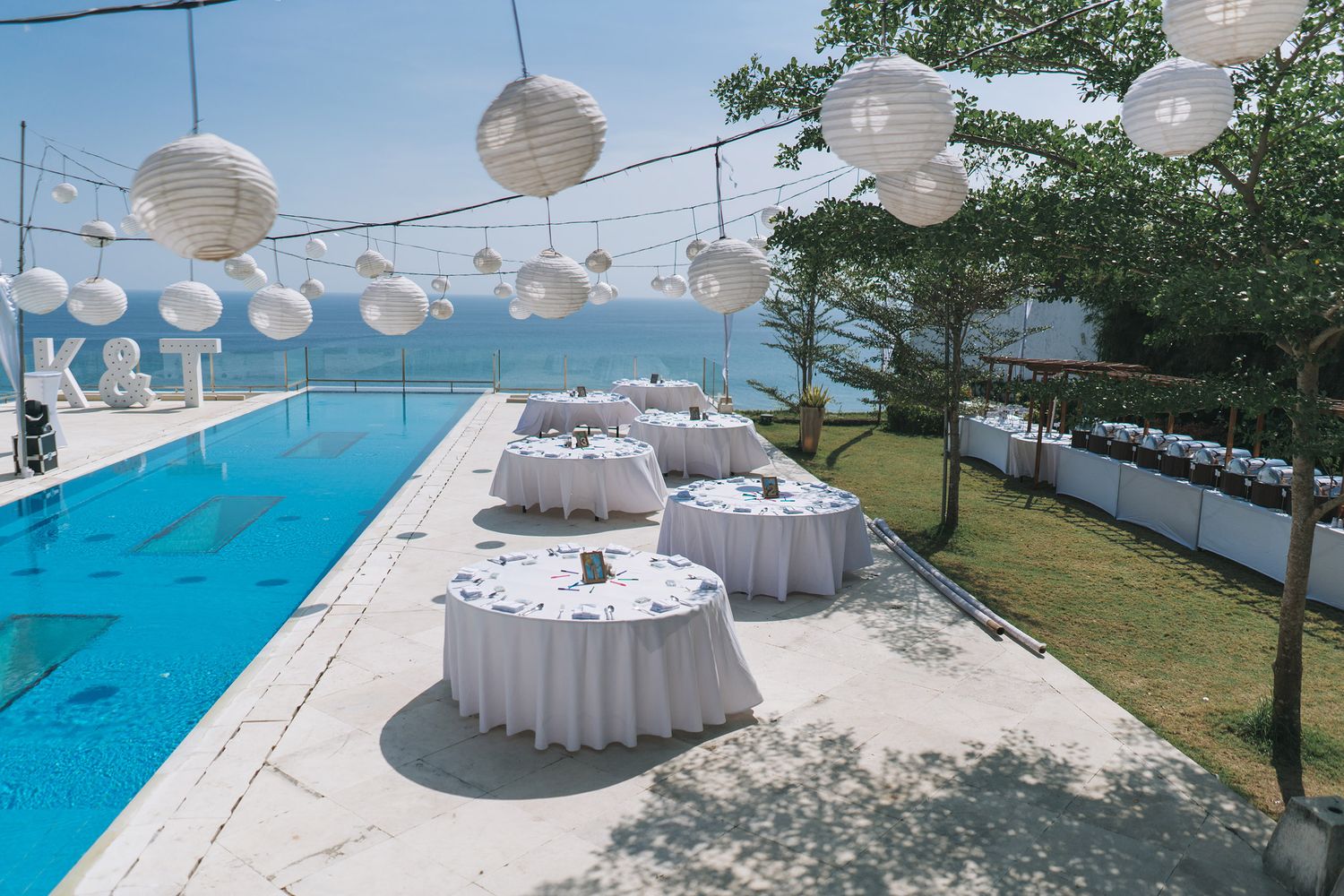 Elegant outdoor wedding reception setup with white lanterns and round tables by swimming pool.