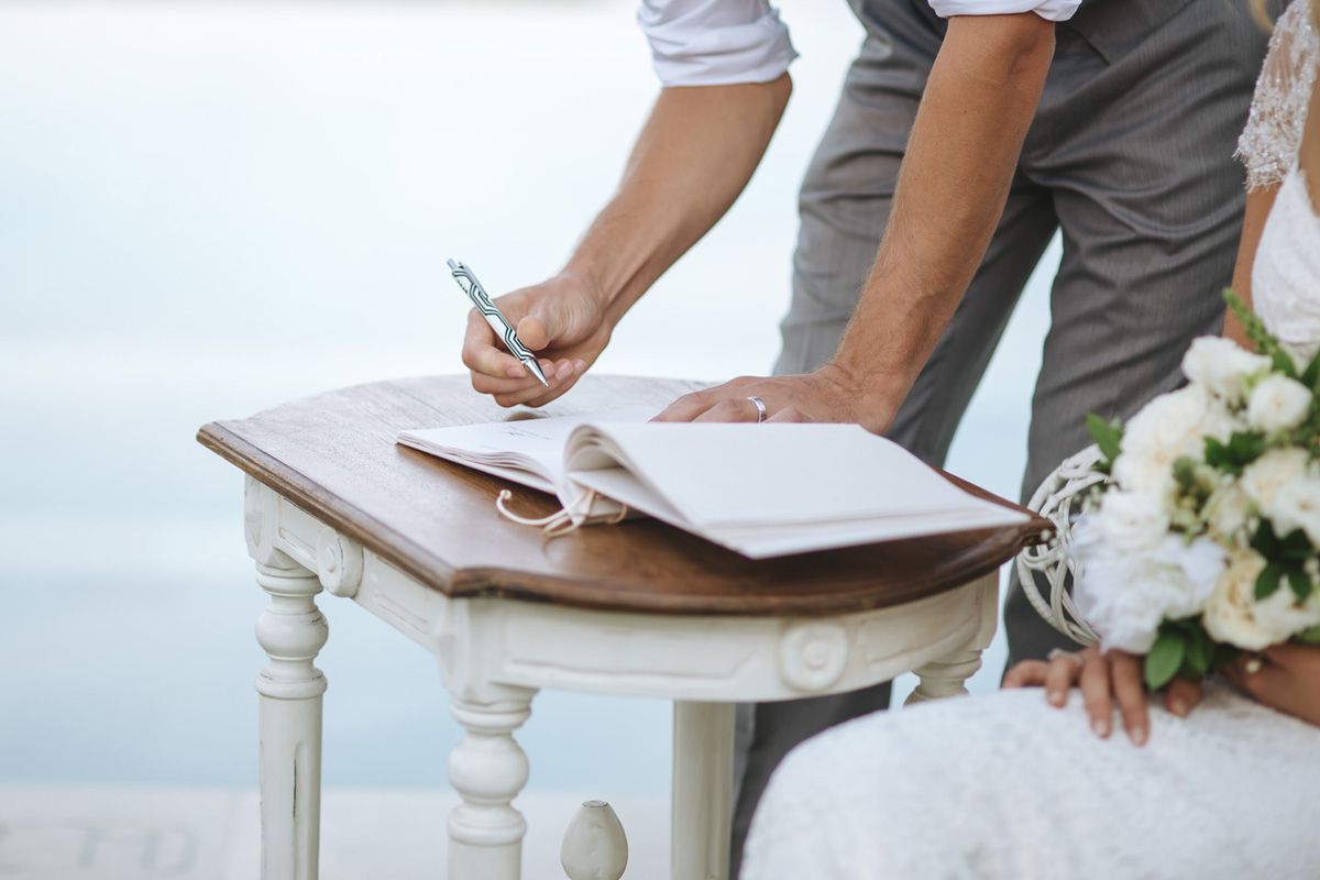 Hands signing marriage documents on white ornate table with wedding rings and bouquet visible.