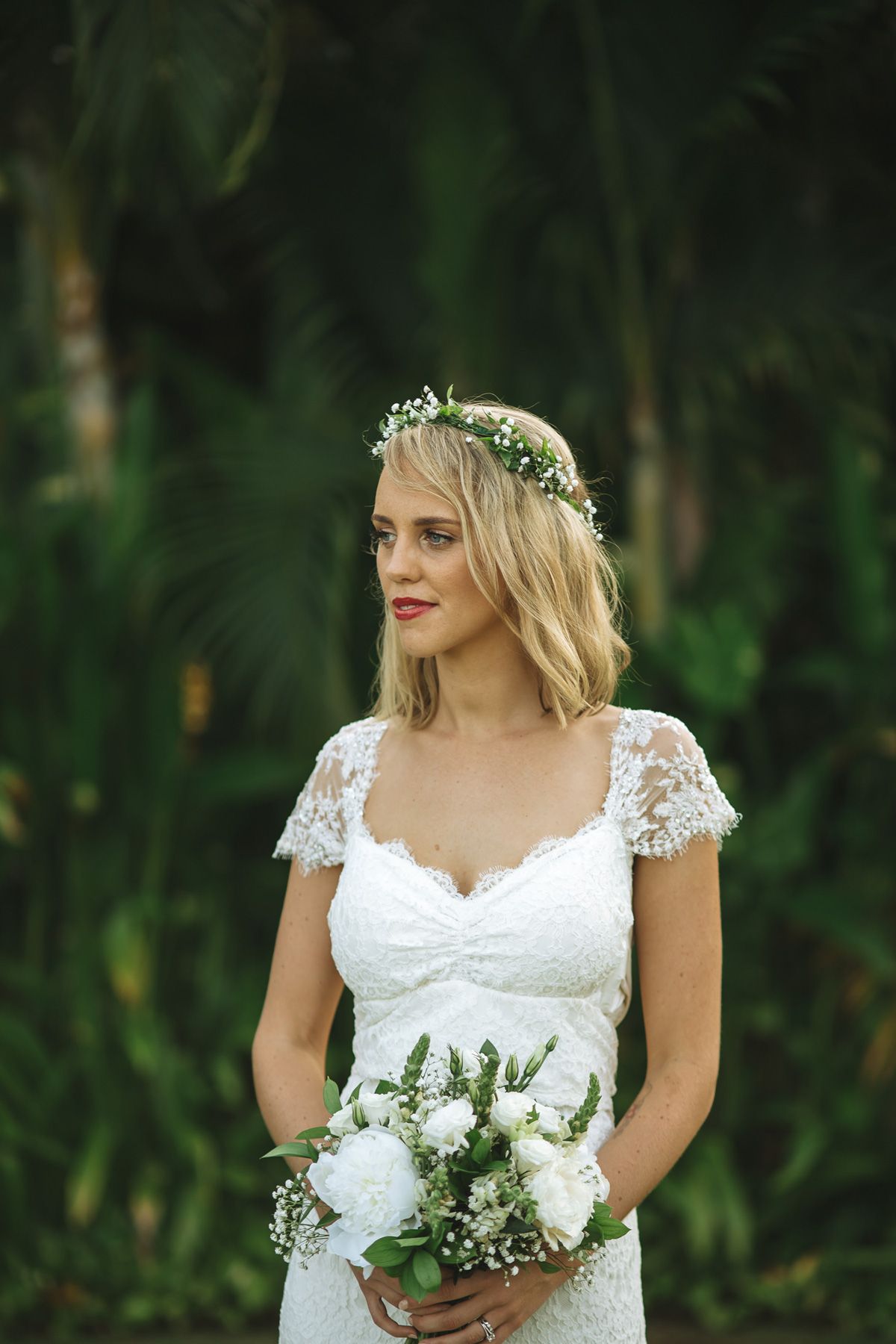 Close-up portraits of bride wearing lace cap sleeve wedding dress and floral crown in natural garden setting.