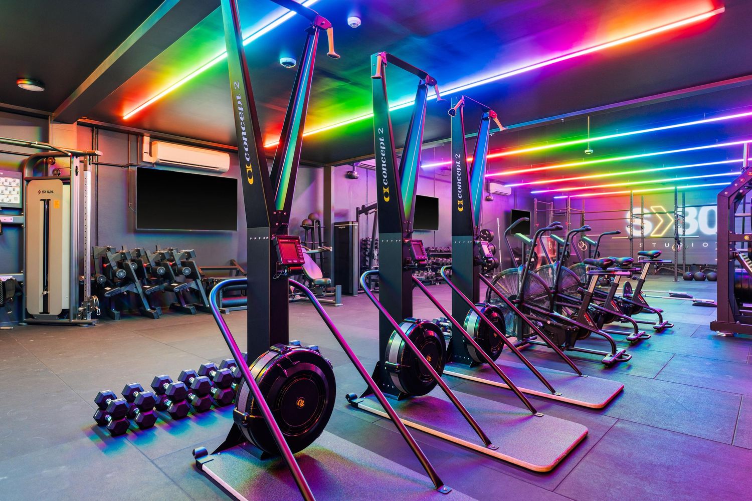 Fitness center with row of exercise bikes illuminated by colorful rainbow LED strips along the ceiling.